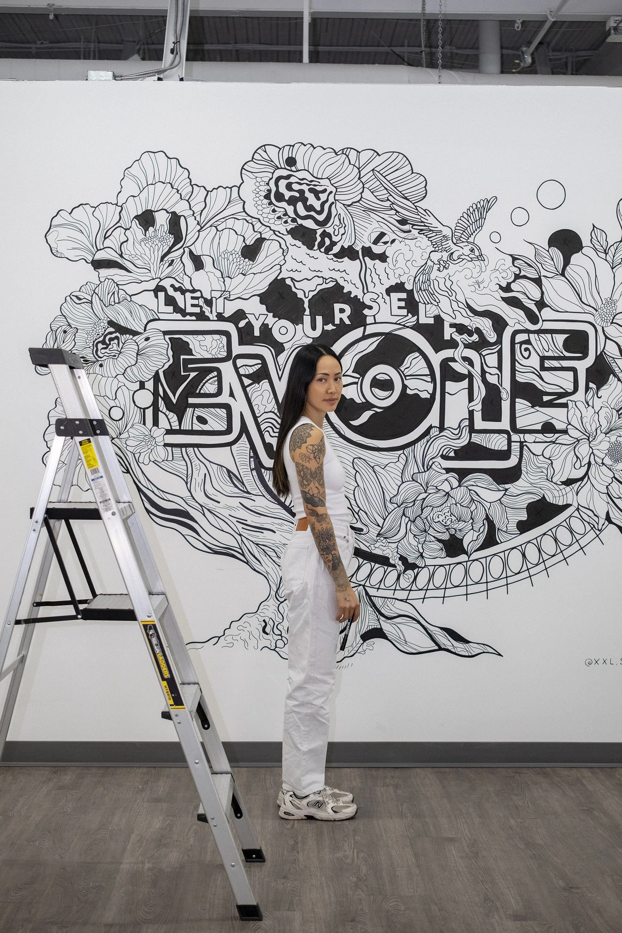 A woman with long dark hair and tattoos on her arms standing next to a ladder in front of a large black and white mural with floral designs and the words "LOVE YOURSELF".