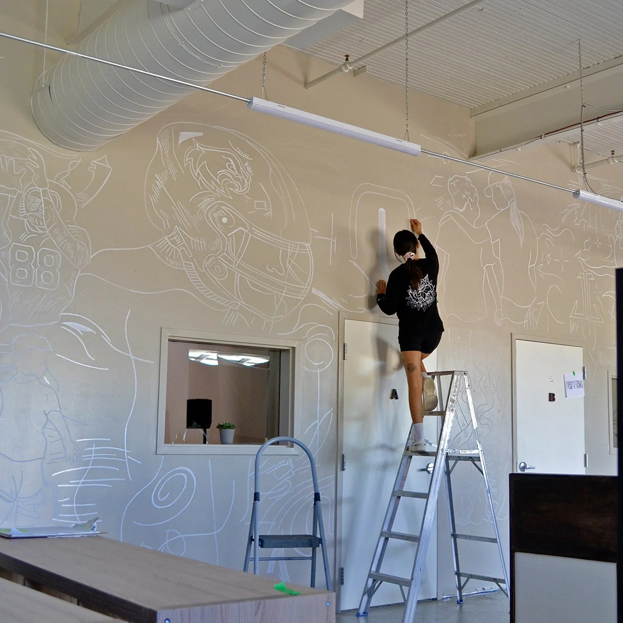 A woman standing on a ladder drawing white chalk sketches of football players and sports scenes on a beige wall.