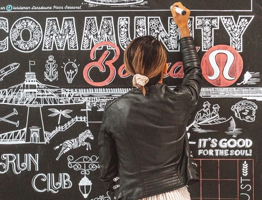 A woman with a ponytail writing on a large blackboard decorated with white chalk drawings and colorful accents, including the word 'COMMUNITY' and the Leo zodiac symbol.