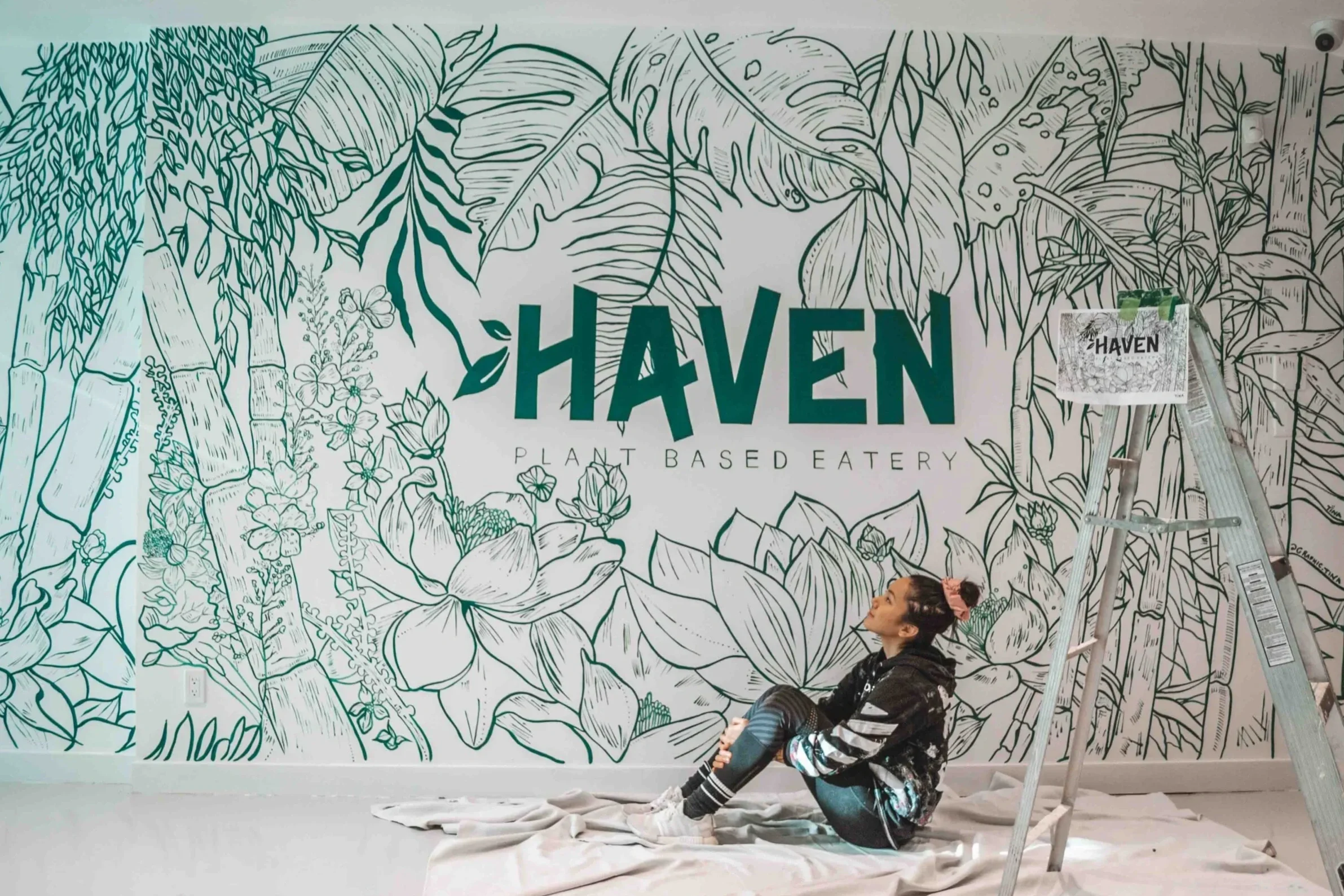 A woman sitting on the floor next to a ladder in front of a large wall mural with the words 'HAVEN Plant Based Eatery' surrounded by botanical illustrations of flowers, leaves, and bamboo.