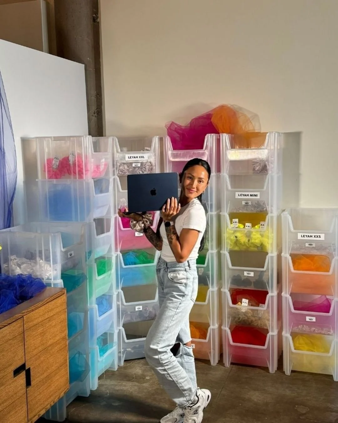 Woman standing in front of clear plastic storage bins filled with colorful fabrics, holding a laptop.