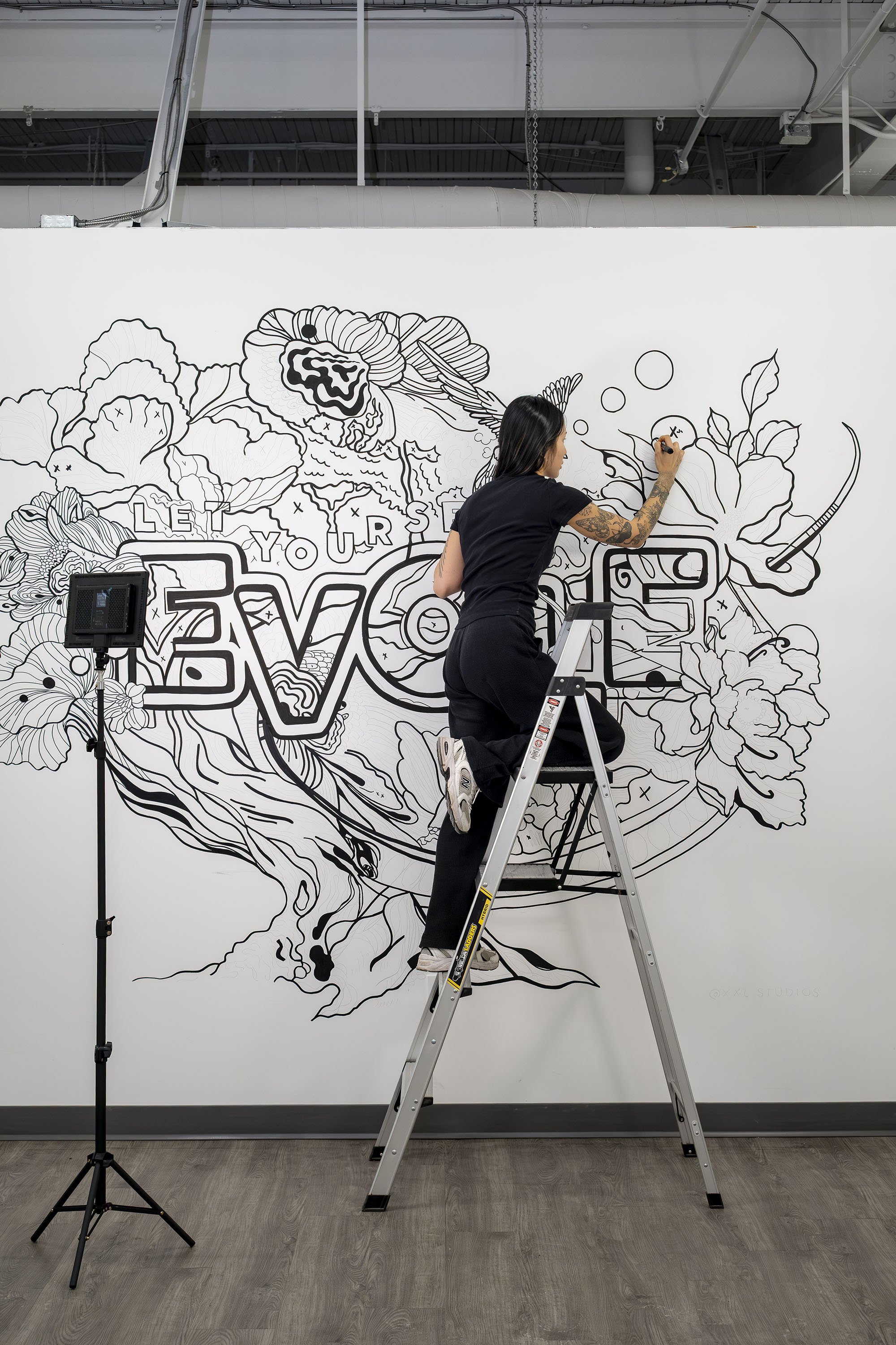 An artist stands on a ladder drawing a mural on a white wall featuring large, stylized text that reads 'LOVE' surrounded by floral designs and abstract shapes.