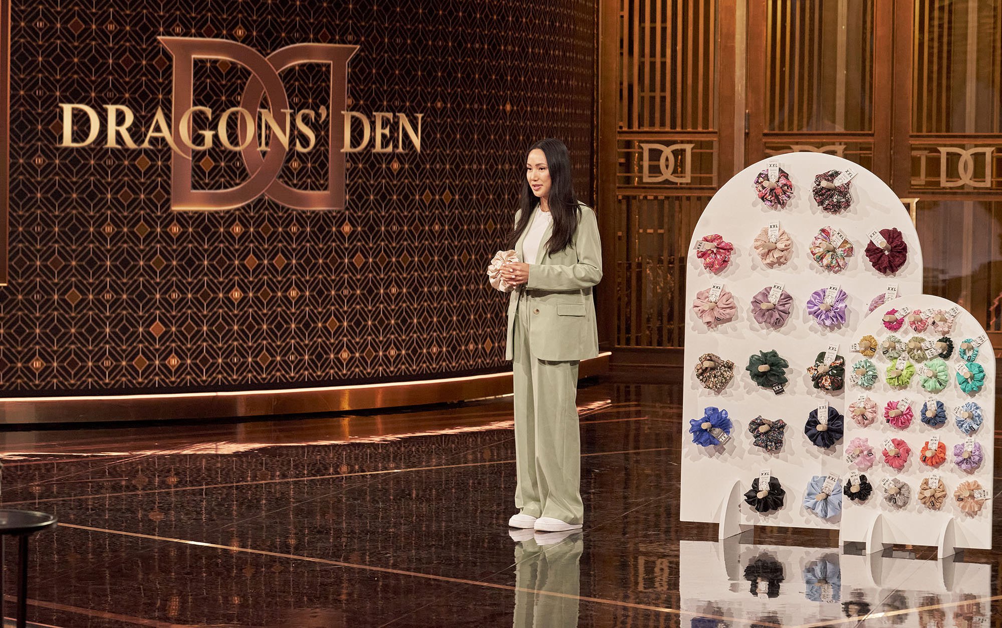 A woman in a beige suit stands in front of a display of colorful scrunchies at an event with a background sign that reads "DRAGON'S DEN."