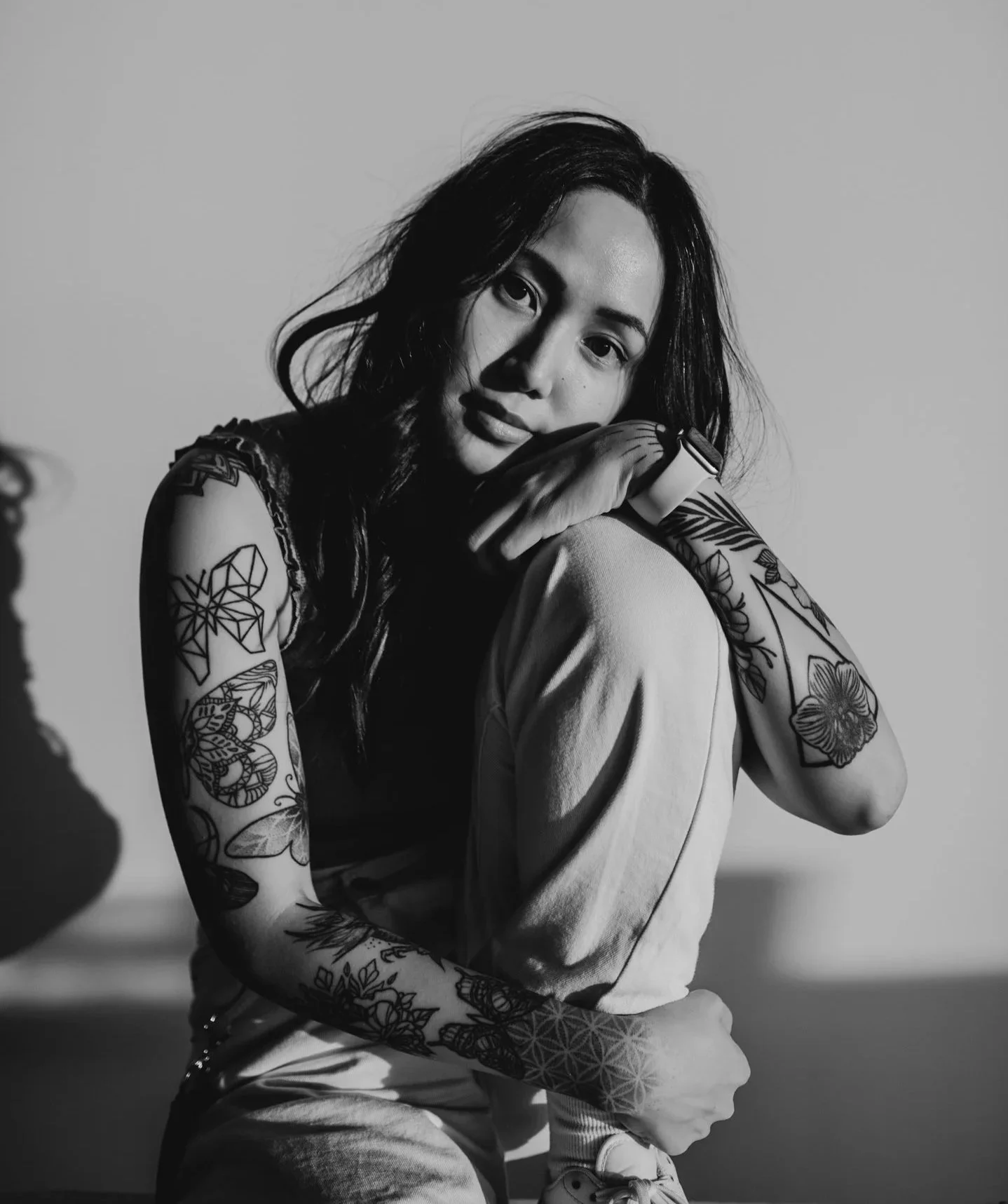 A black and white photo of a woman with long, dark hair and tattoos on her arms, sitting with her knee up and her face resting on her hand, looking at the camera.