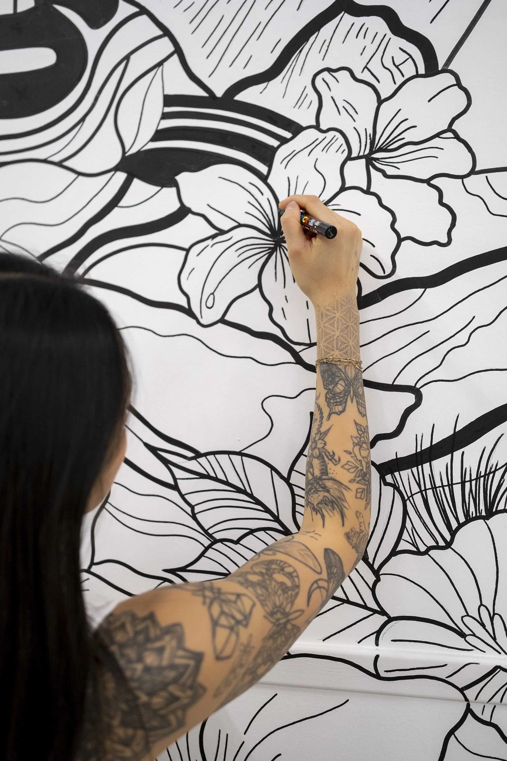 A person with tattoos on their arm drawing a large floral mural on a wall with a black marker.