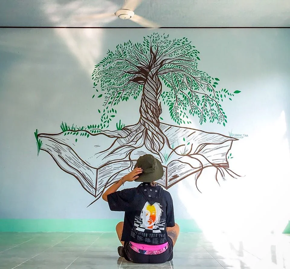 Person sitting on the floor wearing a black t-shirt with a graphic and pink shorts, facing a large wall mural of a tree with green leaves and roots, on a white background.