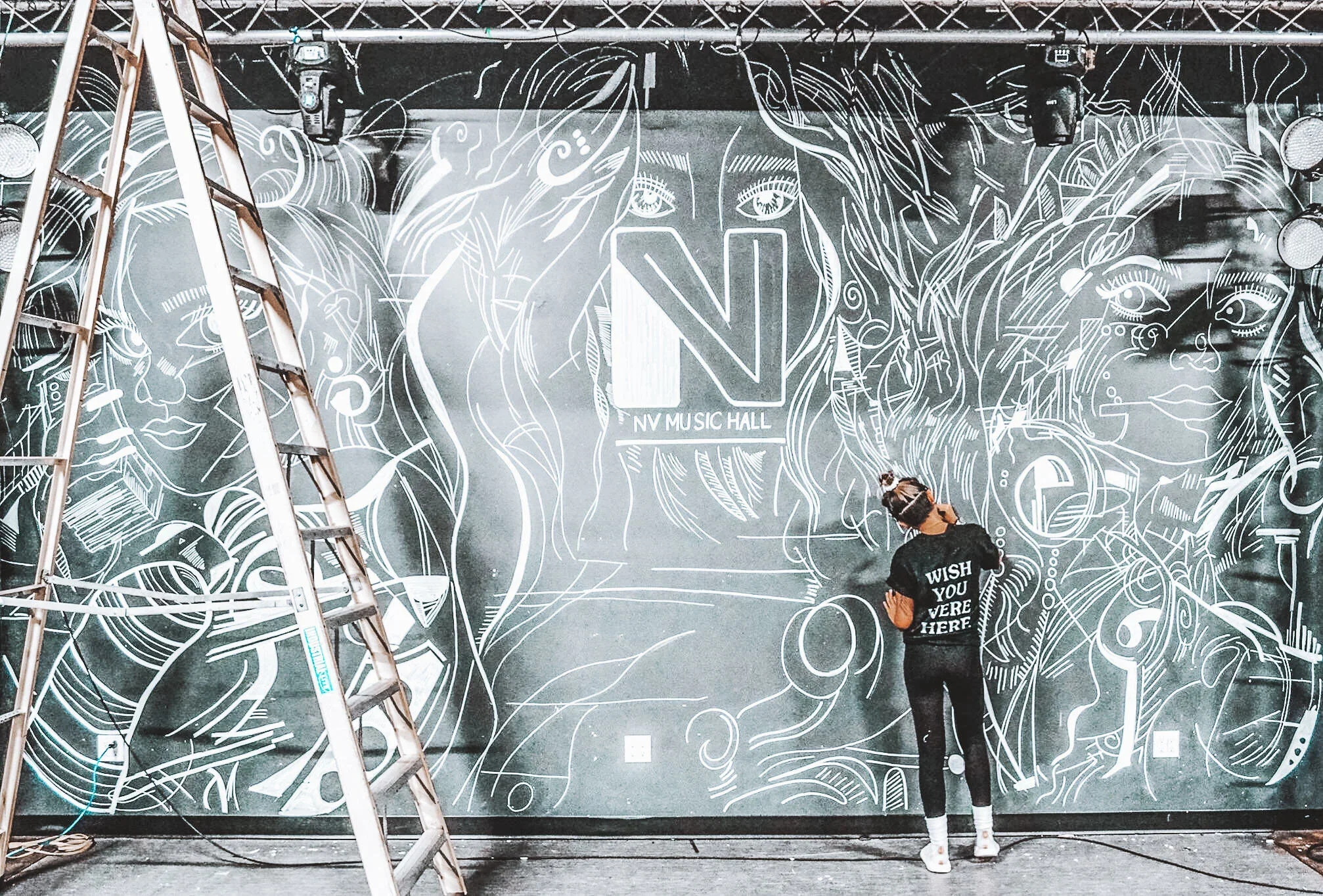 A person is painting a large mural on a wall that features stylized faces and intricate line art. The person is wearing a black shirt that says 'WISH YOU WERE HERE' and white shoes, and is standing in front of the mural. There is a ladder on the left side of the image.