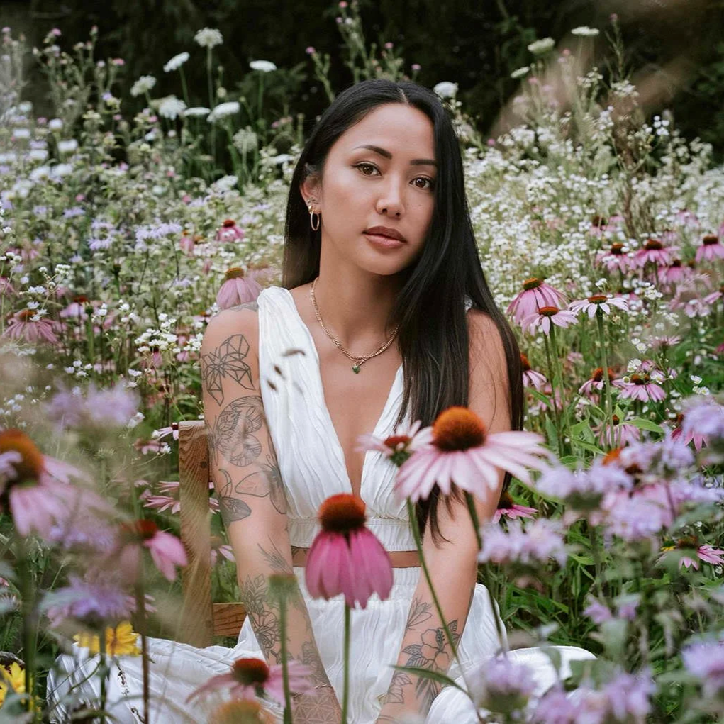 A young woman with long black hair and visible tattoos on her arms sits in a garden filled with pink and purple coneflowers and other wildflowers, wearing a white sleeveless dress and gold jewelry, posing for a photo.