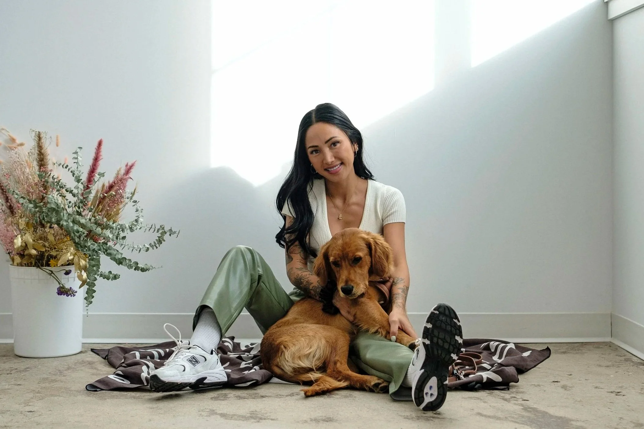 A woman with long black hair, tattoos on her arms, wearing a white top, green pants, and white sneakers, sitting on the floor with a brown puppy, in a minimalist room with a white wall and a potted flower arrangement.