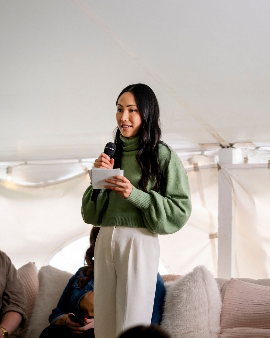A woman with long black hair, wearing a green sweater and white pants, speaking into a microphone while holding notes, inside a tent or large white canopy with cushions on the sofa.