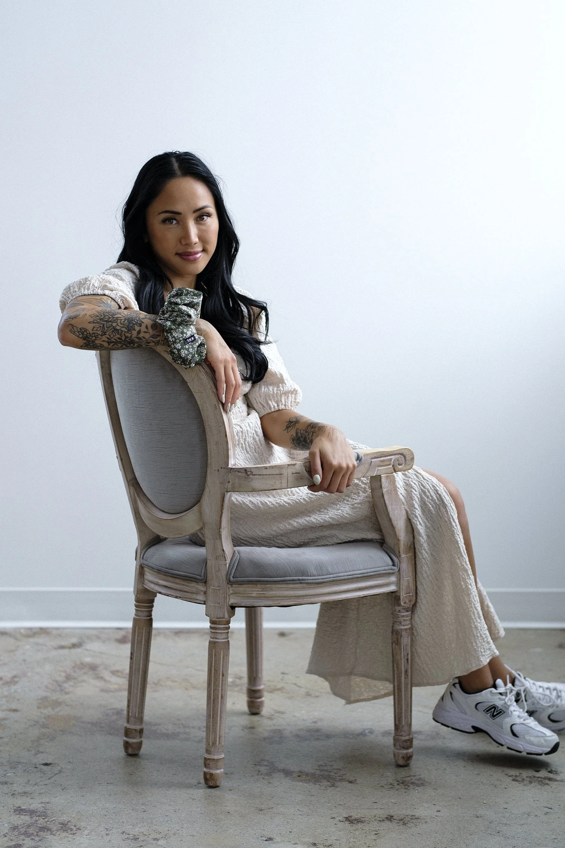 Woman with tattoos wearing a beige dress and sneakers, sitting sideways on a vintage chair with light wood frame and gray upholstery, against a plain white wall.