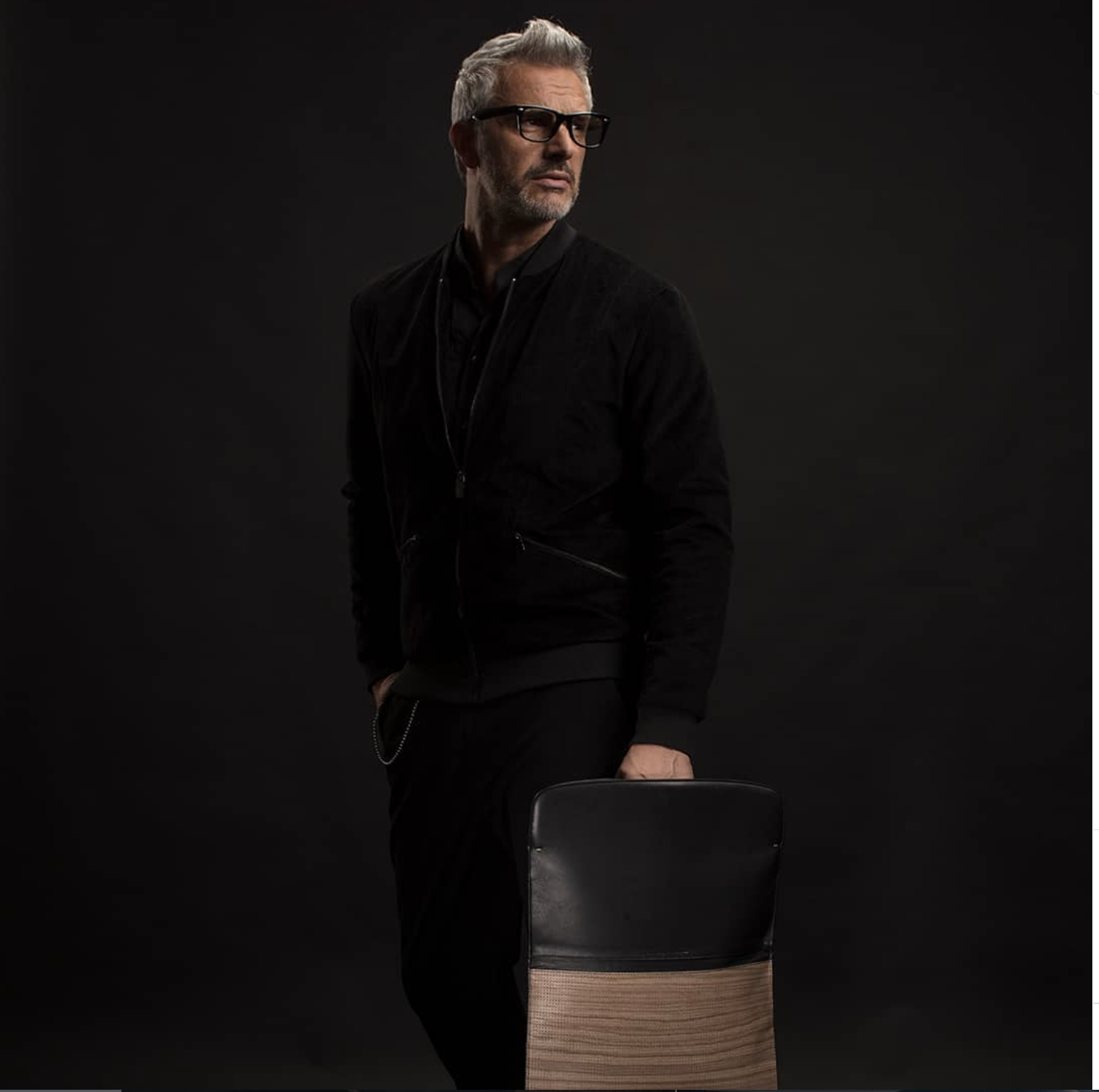 Uomo con capelli grigi e barba, indossa occhiali spessi neri e un giacchino nero, si appoggia a una sedia di fronte a uno sfondo nero. pininfarina folio, advertising, campagna pubblicitaria, strategia social.
