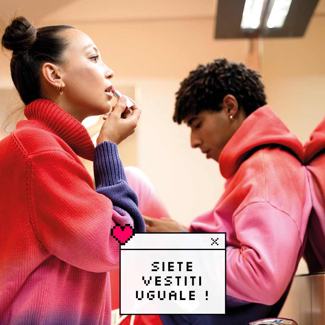 Due giovani indossano cappotti colorati. Una ragazza sta mettendo il rossetto, mentre un ragazzo guarda distrattamente. Un messaggio sullo sfondo dice "Siete vestiti uguale!" con un'icona a forma di cuore pixelato.