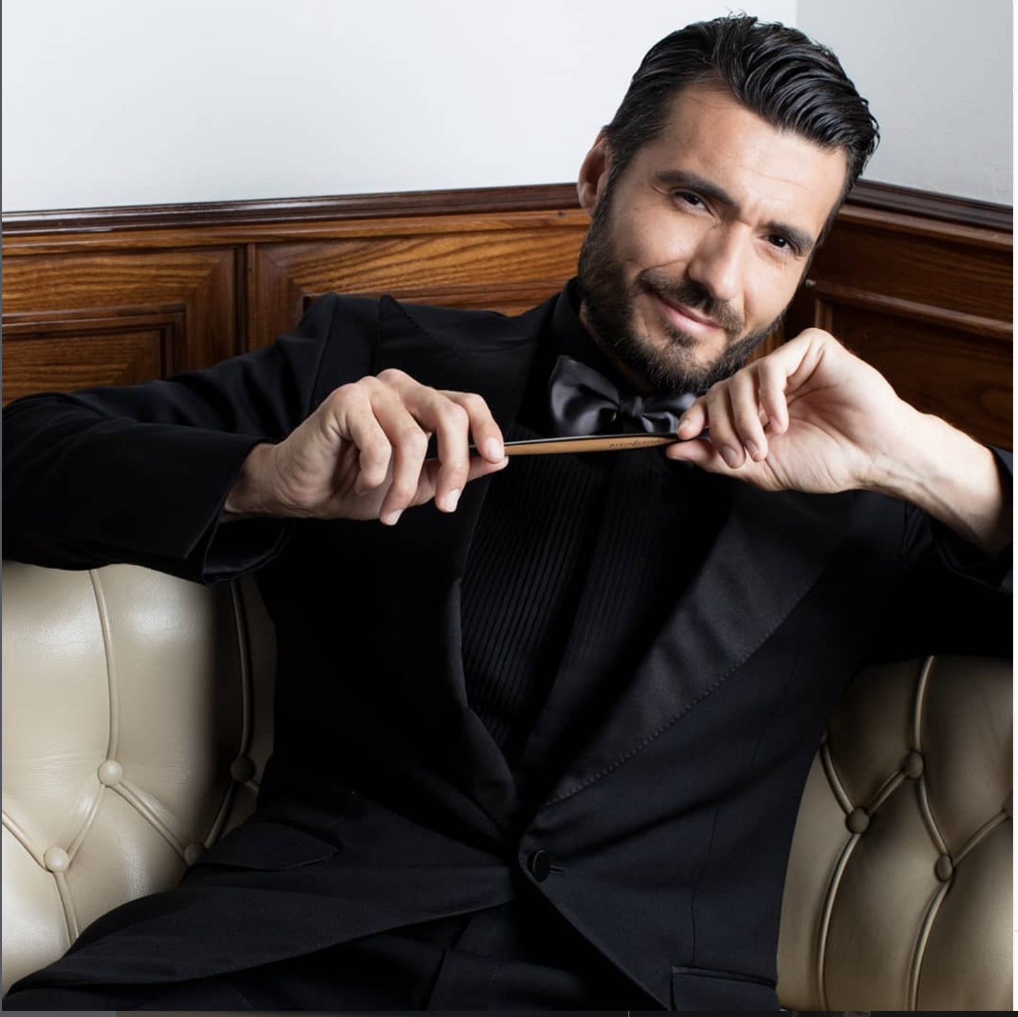 Uomo elegante con smoking nero e papillon, seduto su un divano imbottito, che sorride e tiene una bacchetta da direttore d'orchestra, con pannello di legno sullo sfondo. stilo pininfarina cambiano, campagna pubblicitaria, advertising, strategia 
