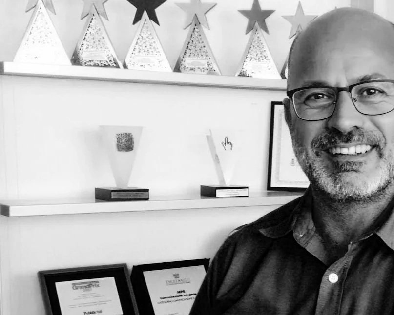 Uomo con occhiali e barba che sorride in un ambiente decorato con stelle e premi su scaffali.