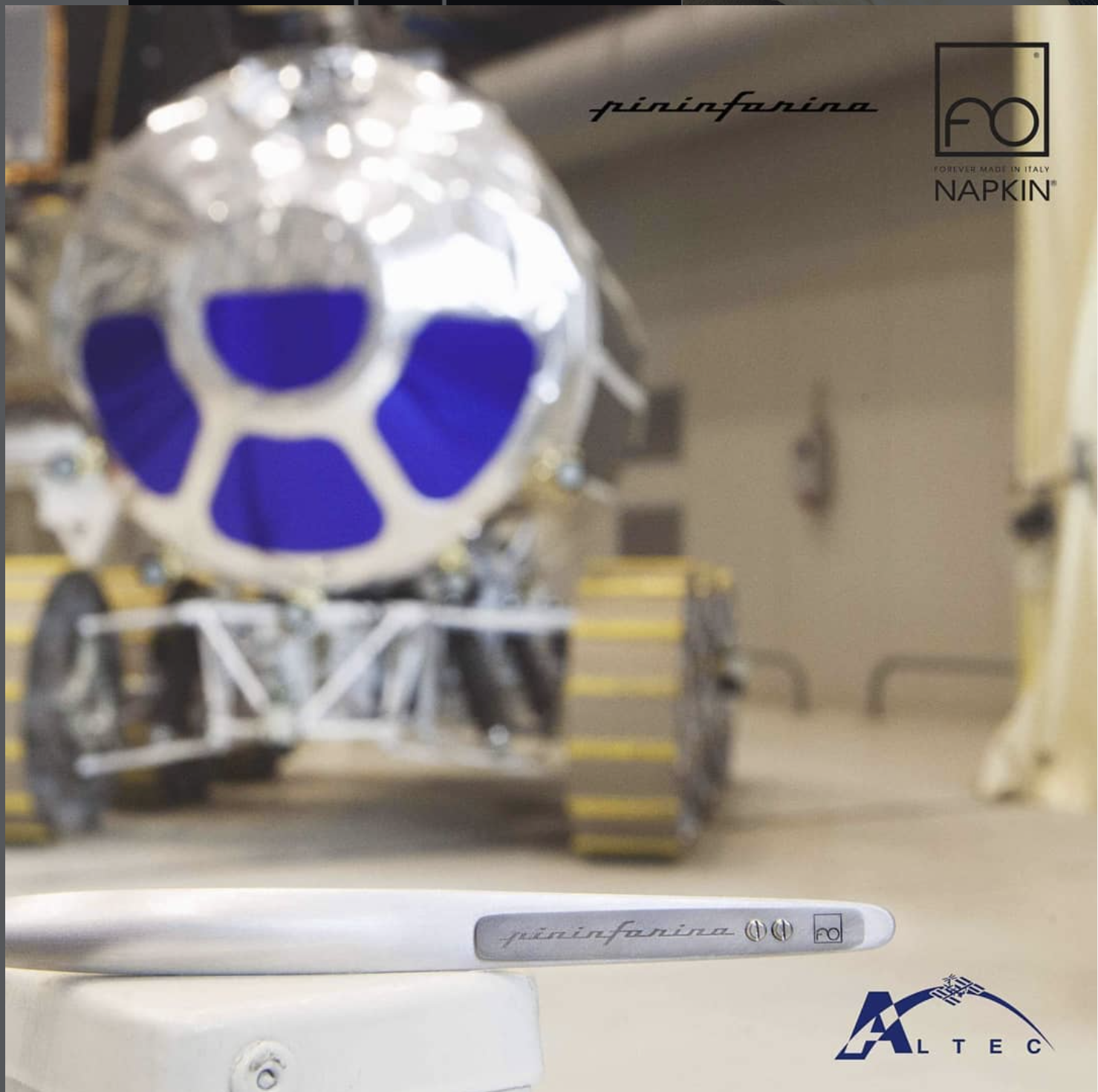Una penna bianca con il marchio 'pininfarina' space, Sullo sfondo, un modello di una navicella spaziale con la parte frontale blu e dettagli metallici, e un pavimento di colore chiaro e pareti di colore beige in un ambiente industriale. altec, ISS,