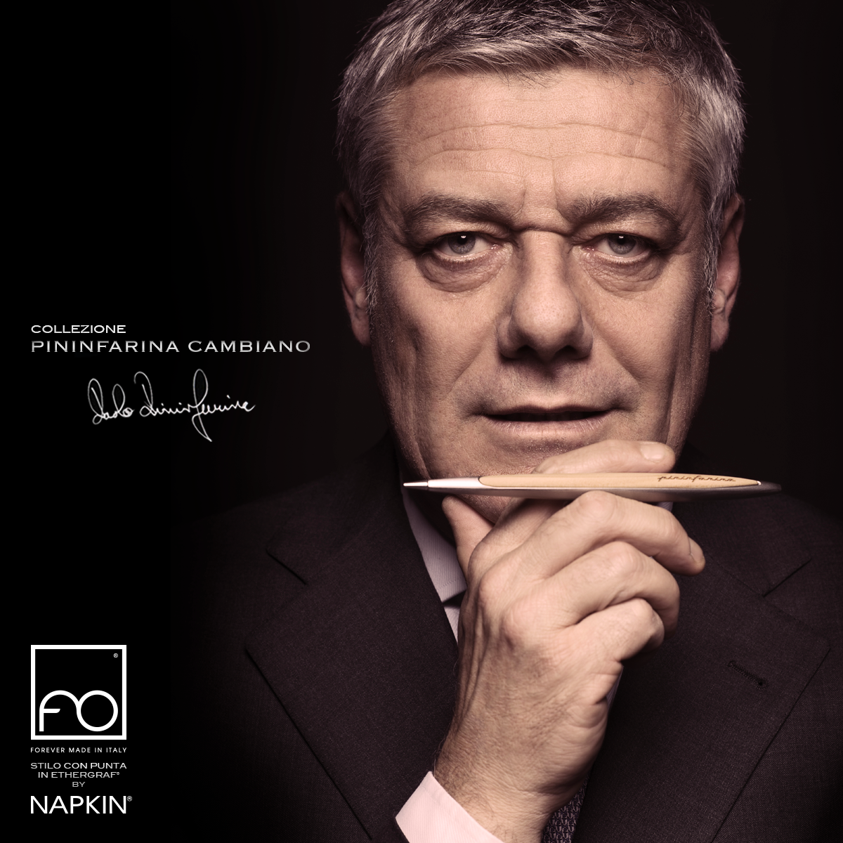 Ritratto di un uomo maturo con capelli grigi, che indossa un completo scuro, tiene una penna vicino al volto e guarda intensamente verso l'obiettivo. Sul lato sinistro dell'immagine, ci sono testo e firma che promuovono la collezione Pininfarina Cambiano di napkin.
