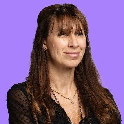 Una donna con capelli castani e occhi chiari, indossa un top nero con dettagli in pizzo e collane, sfondo viola.