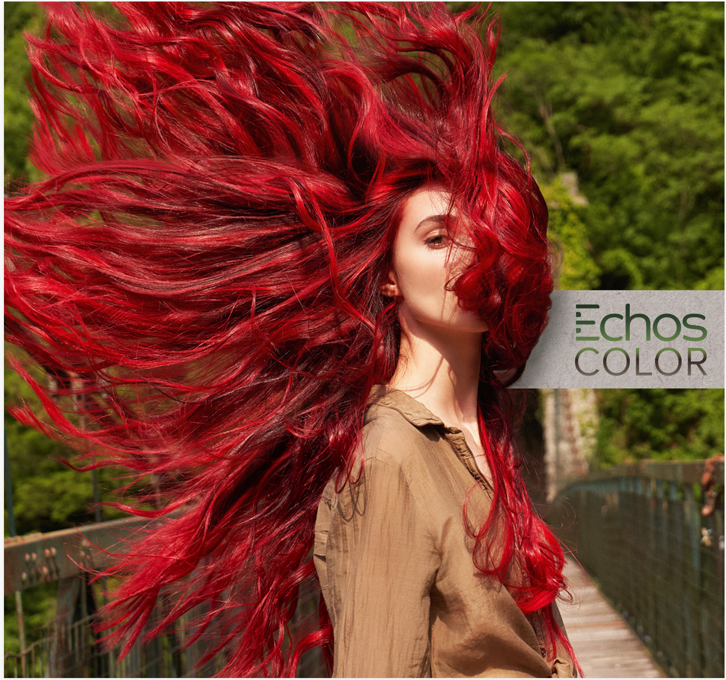 Donna con capelli ricci, rossi e fluenti che sventolano all'aria, su un ponte in un ambiente naturale con alberi verdi, occhio a metà viso coperto, vestita con camicia marrone.