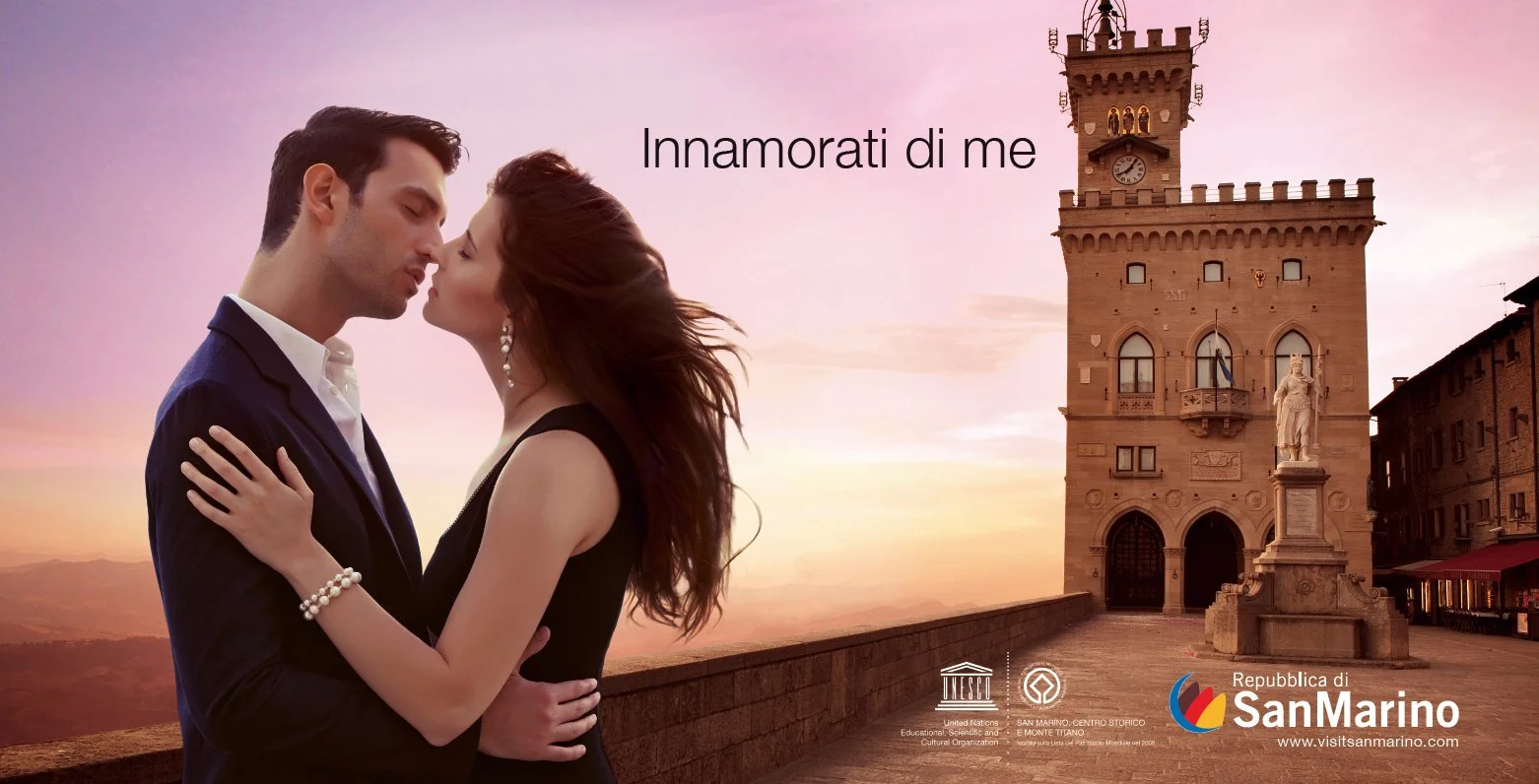 Due persone innamorate si baciano davanti a una torre storica al tramonto, con il testo 'Innamorati di me' sopra. In basso a destra ci sono i loghi dell'UNESCO, San Marino e il sito visitsanmarino.com.