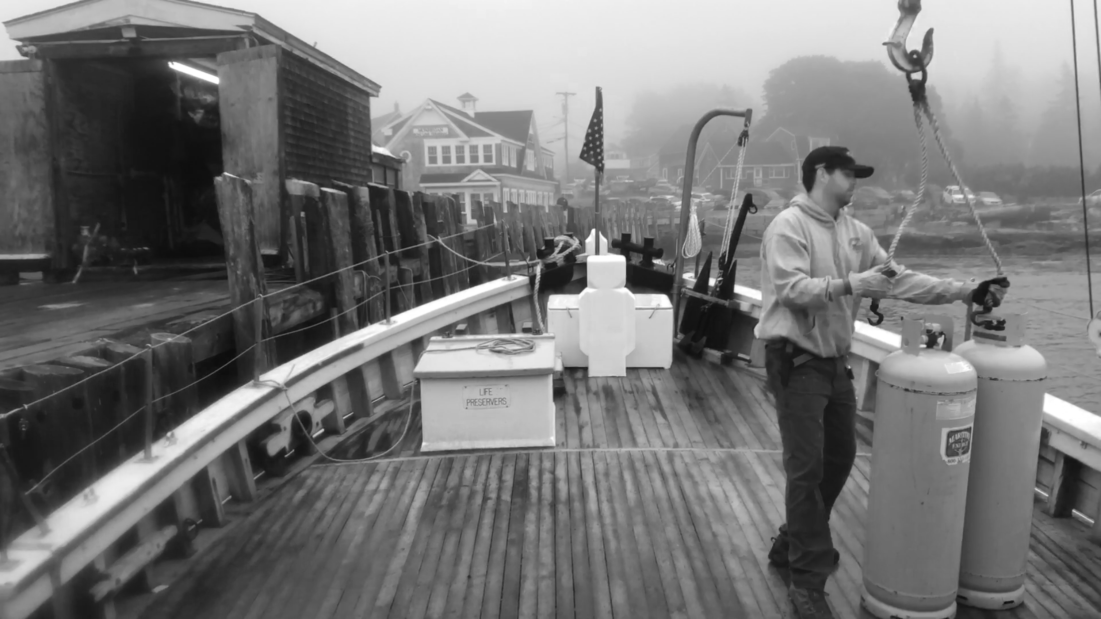 A person on a boat, in foggy weather, holding a rope attached to propane tanks. The boat has fishing gear and an American flag, with houses visible in the background.