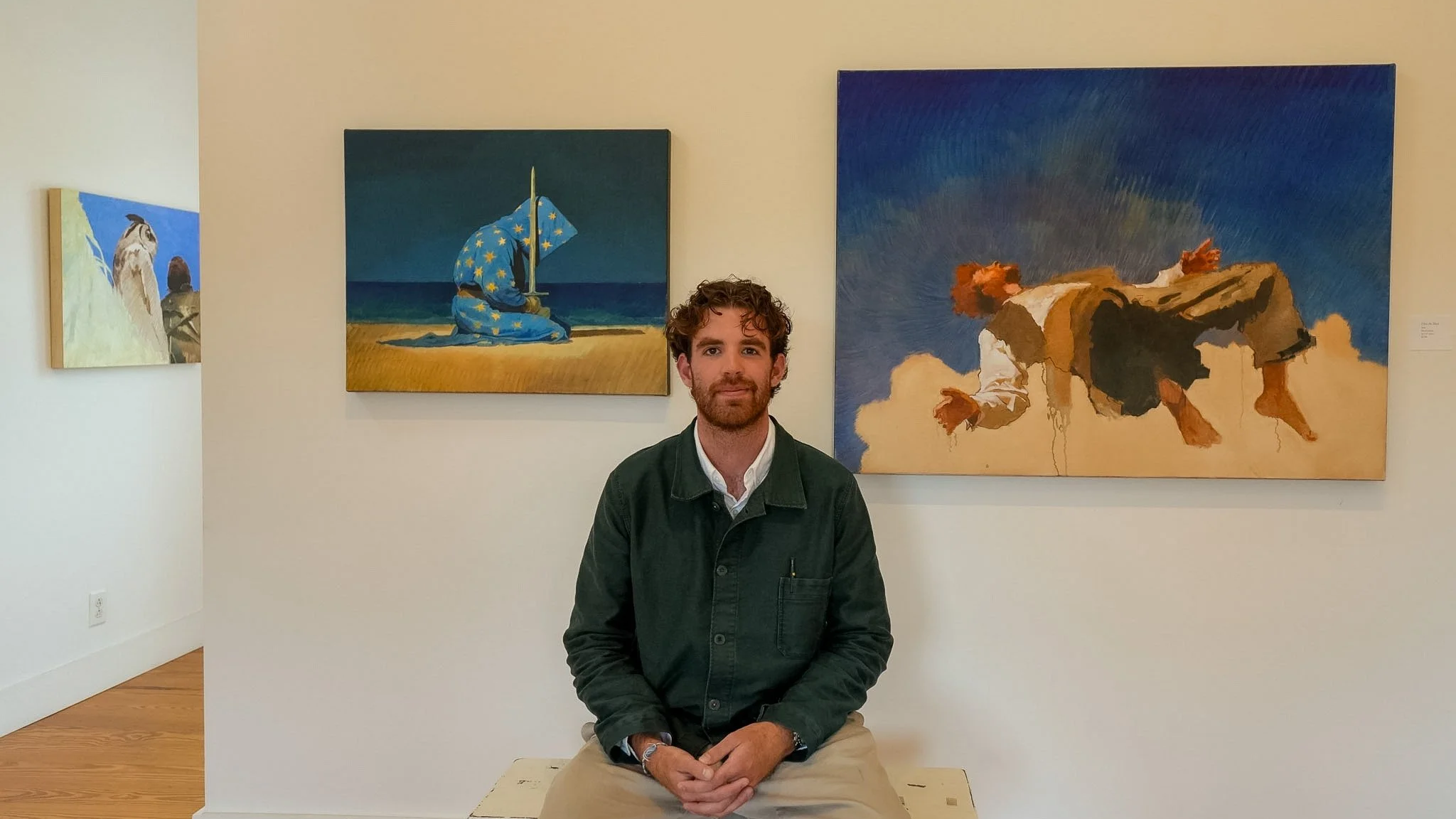 A man with curly hair and beard sitting in front of three paintings in an art gallery.