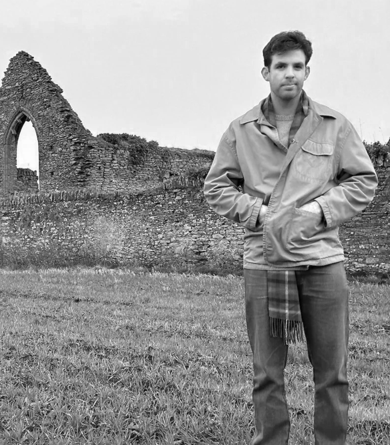 A young man standing outdoors in front of an ancient stone ruin, wearing a jacket and scarf, with hands in pockets, in black and white photography.