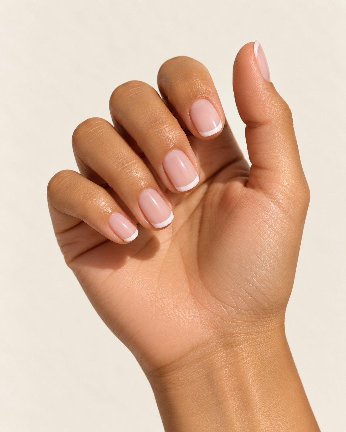 Close-up of a hand with short, glossy pink French manicure nails against a beige background.