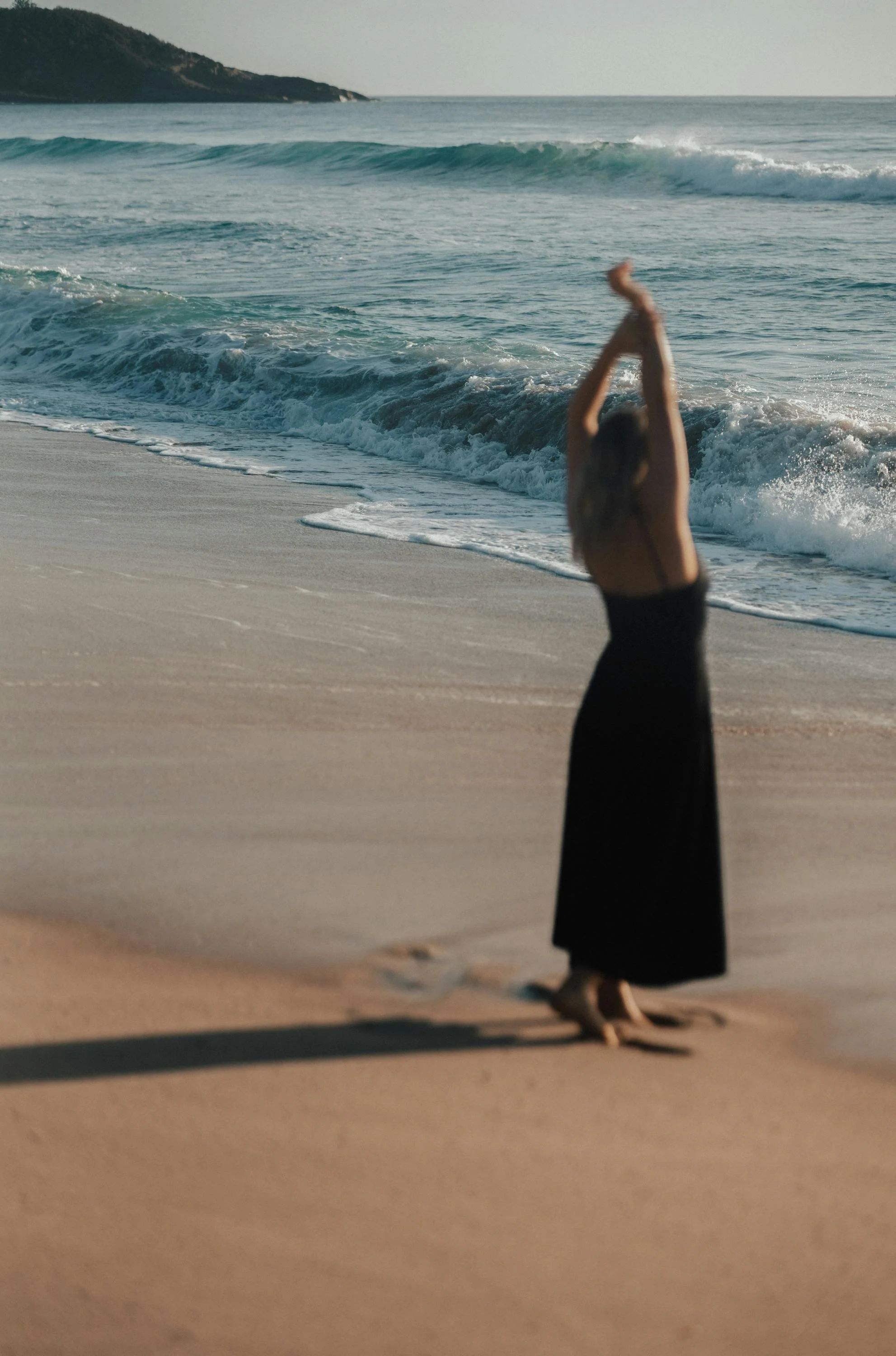 A woman in a black dress practicing yoga in a handstand on a sandy beach near the ocean, with waves and a distant landform in the background.