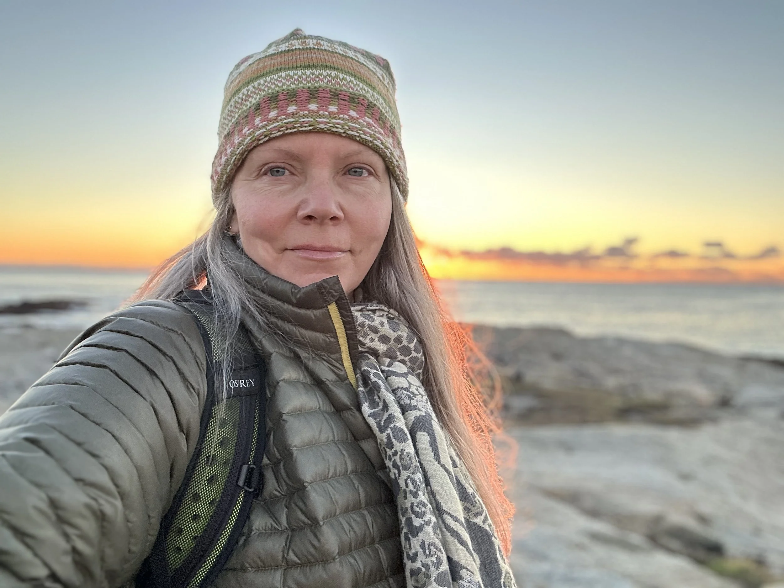 A woman taking a selfie on the beach at sunset, wearing a knitted hat, a puffer jacket, and a scarf, with rocks and the ocean in the background.