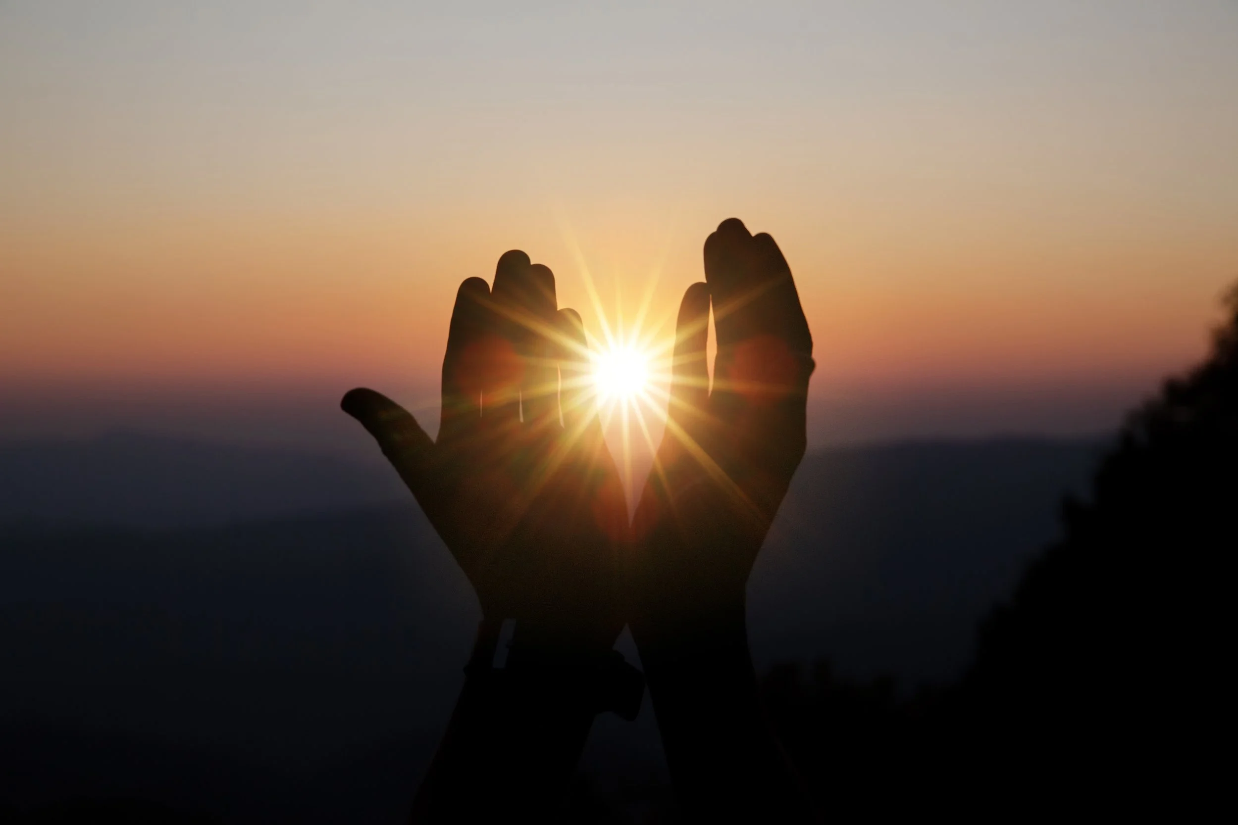 Two hands forming a heart shape around the setting sun during sunset, with a sky in gradient colors from purple to orange.