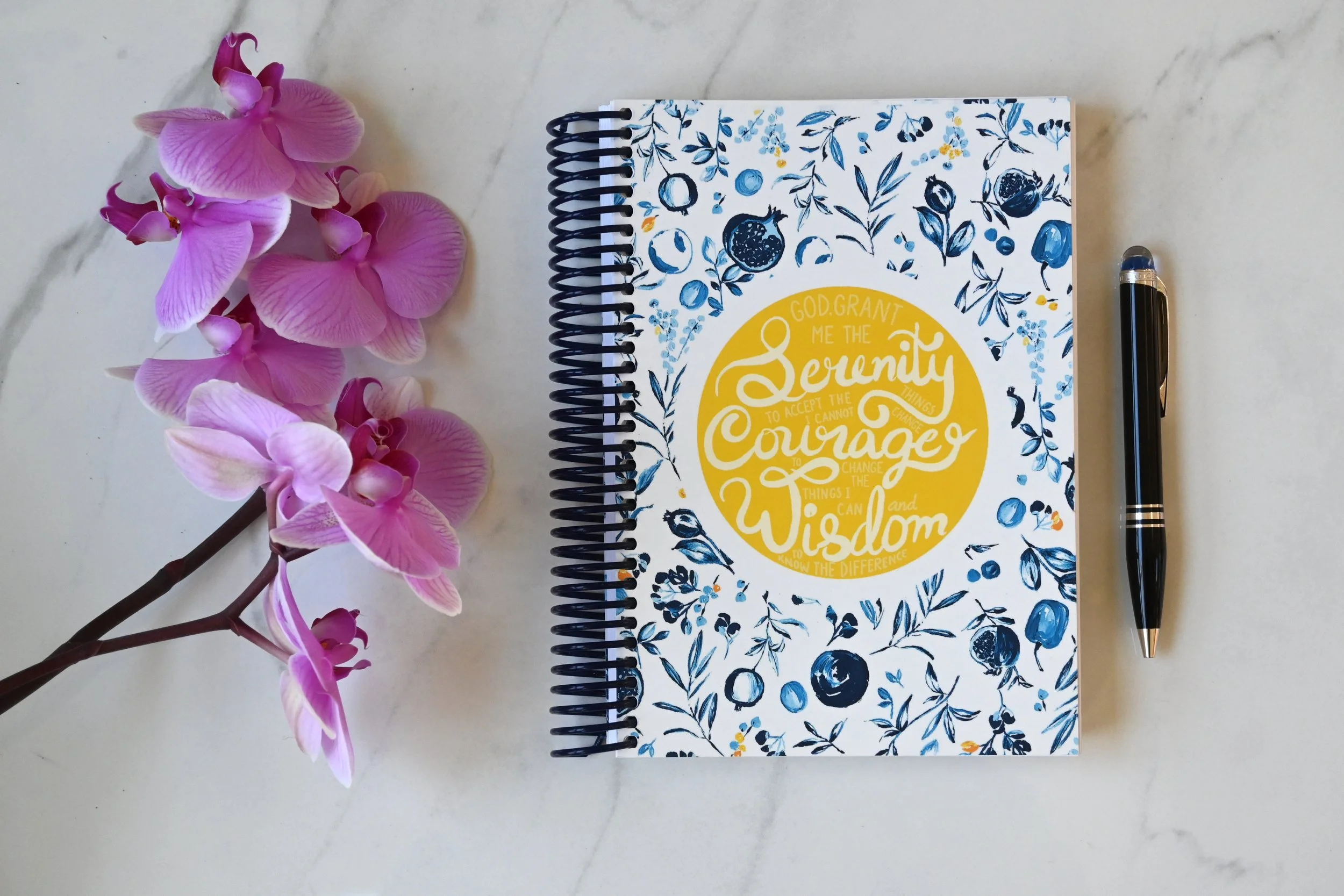 Open spiral notebook with floral cover design, black pen, pink orchids on white marble surface.