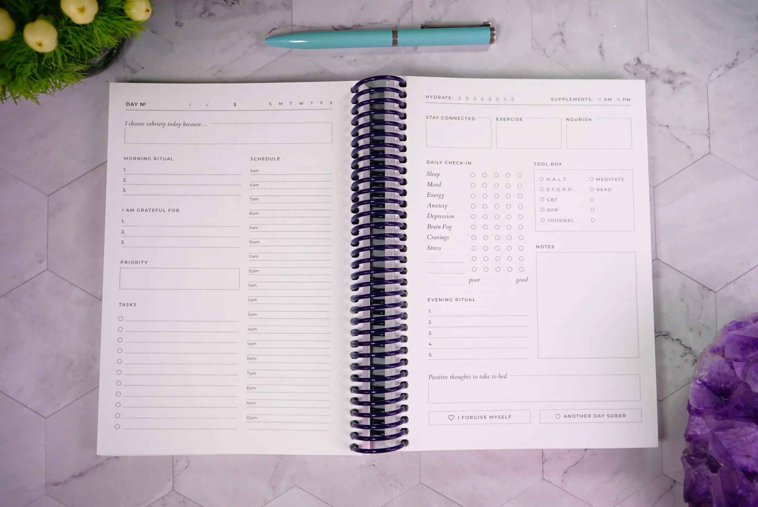 Open spiral planner or journal on marble surface, with a light blue pen above it, and a purple amethyst crystal on the bottom right corner. The pages contain daily and evening rituals, schedule, tasks, and wellness tracking sections.
