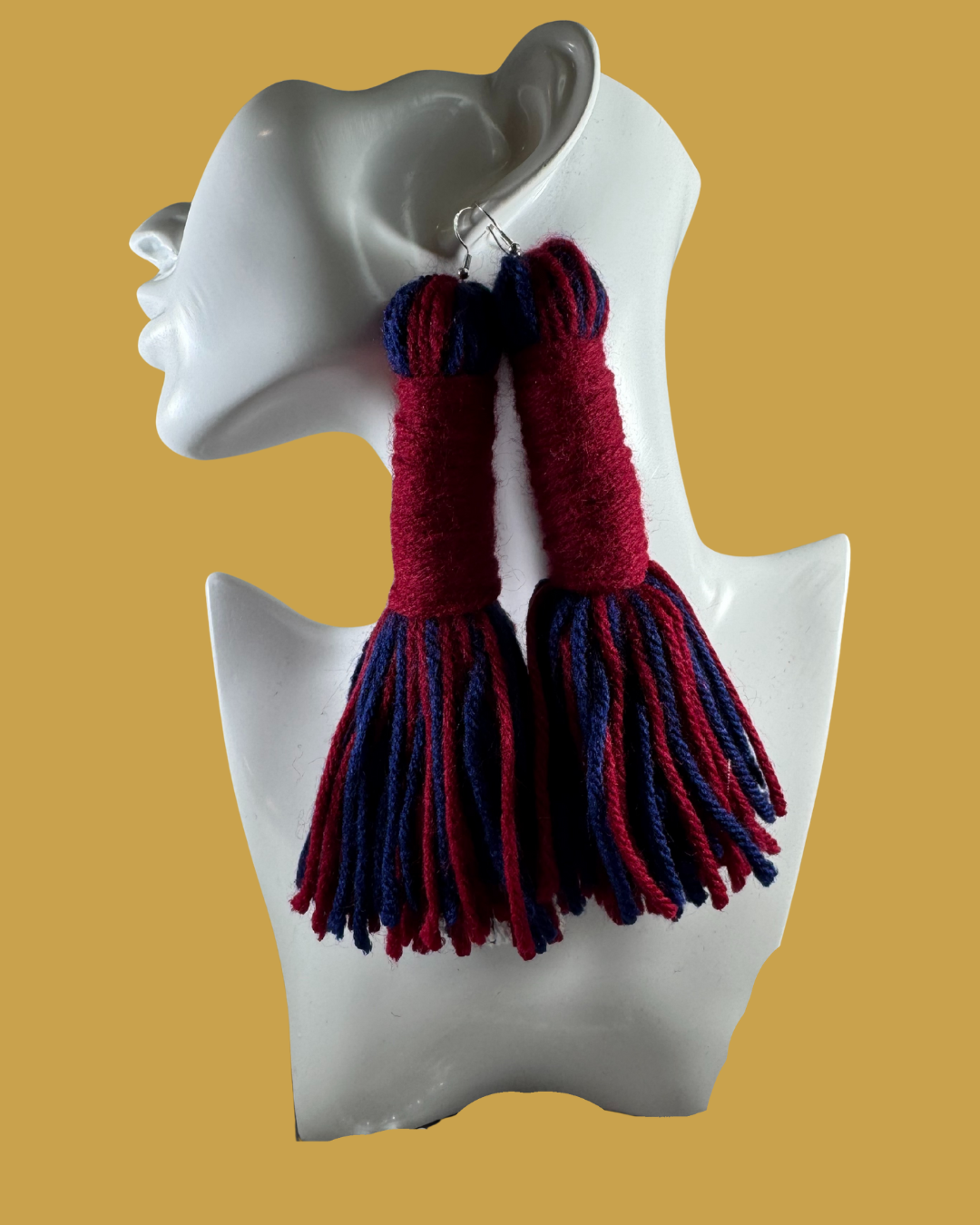 A white mannequin bust displays a pair of handmade woolen earrings and fingerless gloves with a blue and red striped pattern. The gloves have long fringes hanging from the wrists.