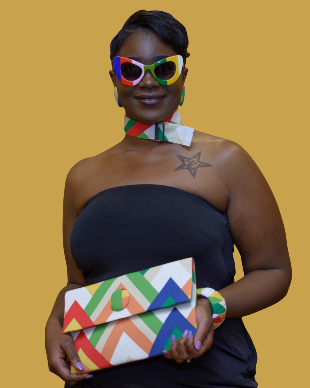 A woman smiling, wearing colorful sunglasses and a matching neck scarf with a black strapless top, holding a multicolored geometric-patterned clutch purse, standing against a plain yellow background.