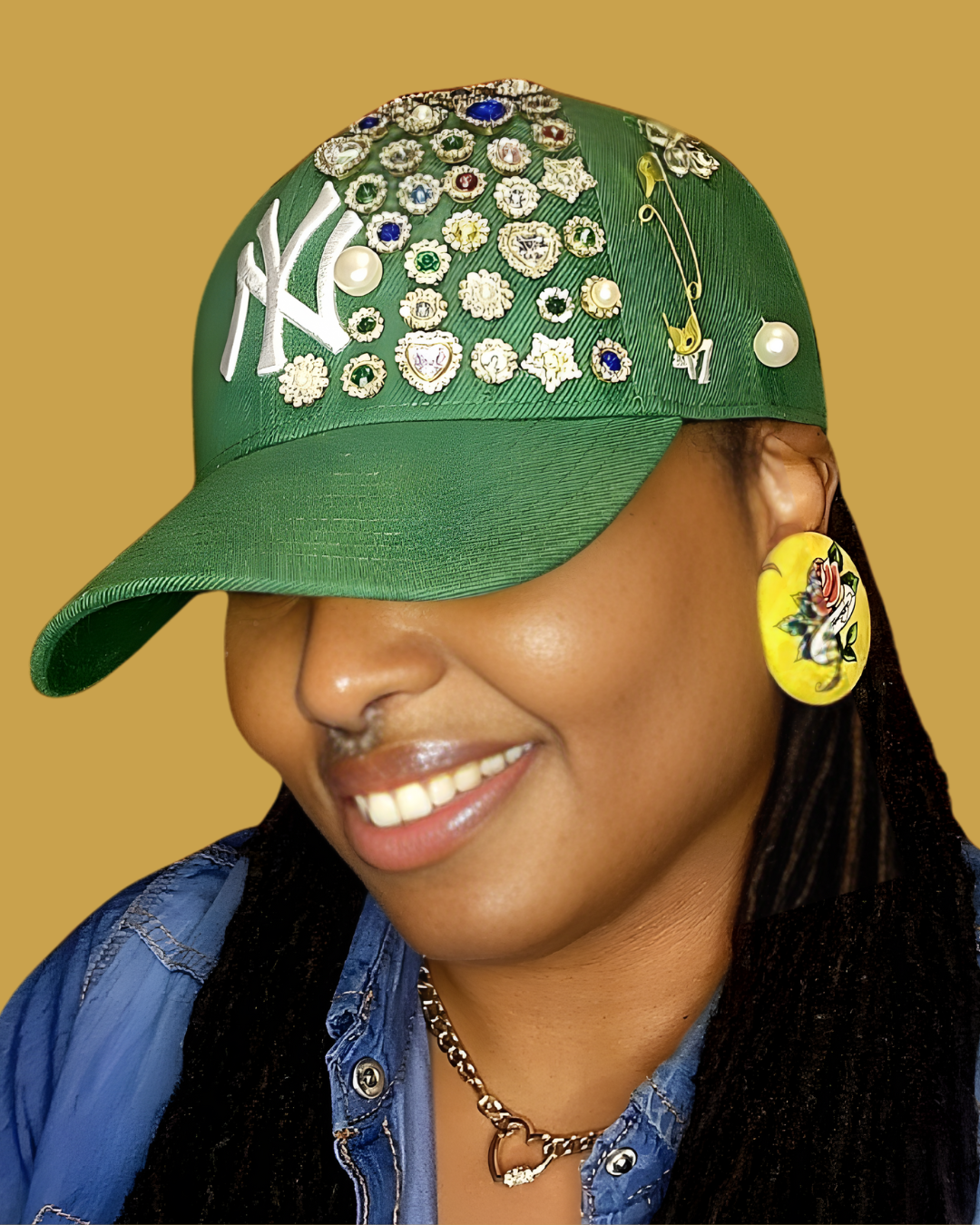 A woman wearing a green baseball cap decorated with jewelry, pearls, and a clock face, with a large yellow earring and a blue denim shirt, smiling against a plain background.