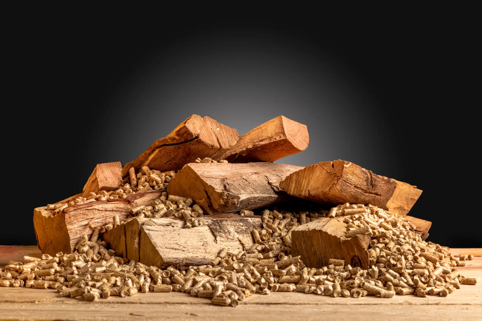 A pile of firewood on a wooden surface with small pellets surrounding it, against a dark background.