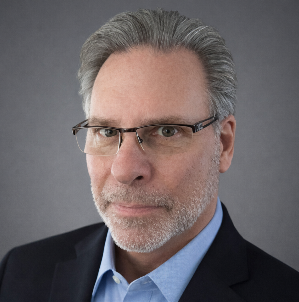 A middle-aged man with gray hair, glasses, and a beard wearing a navy blazer and light blue shirt against a gray background.