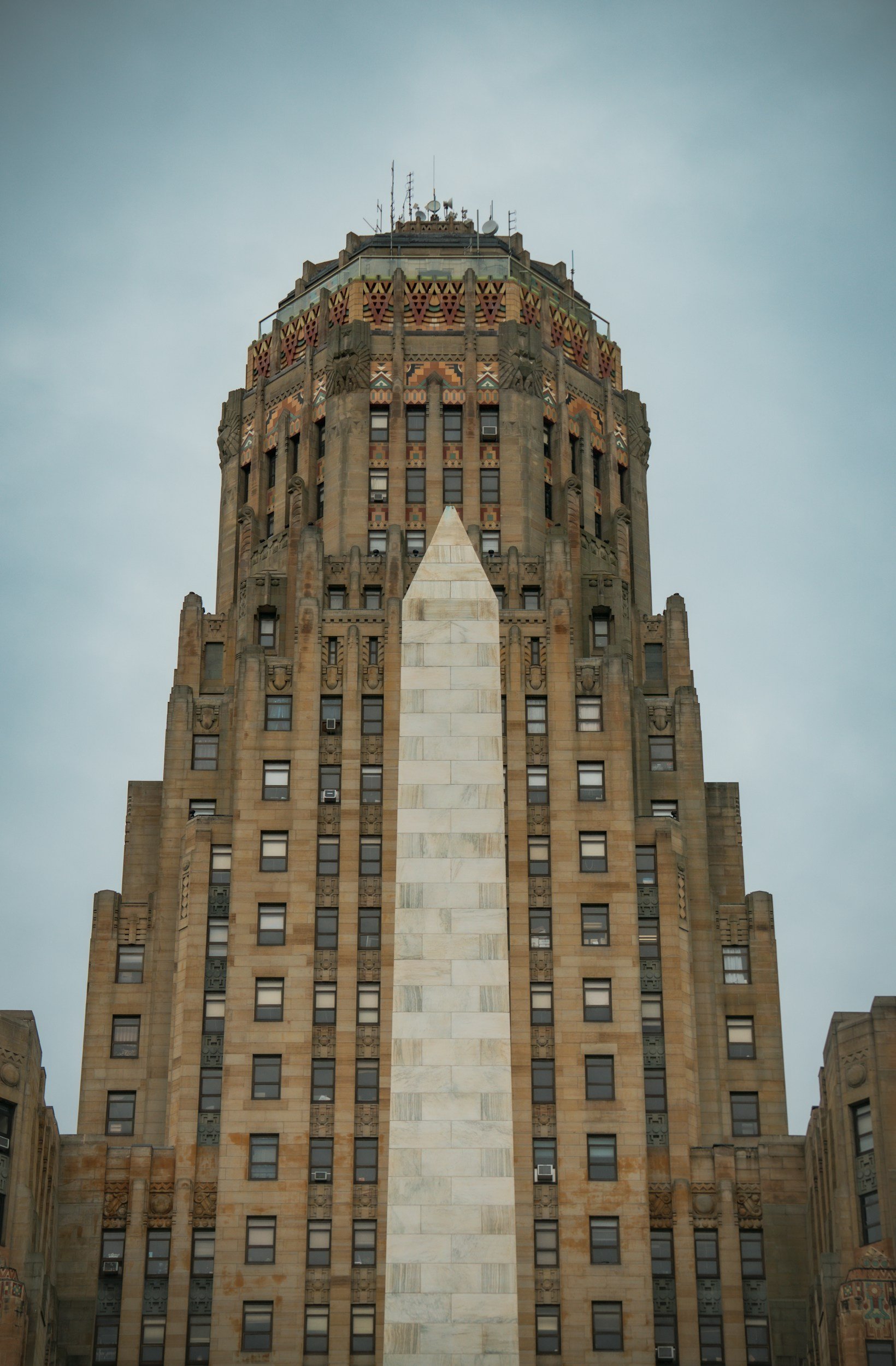 A tall, historic skyscraper with Art Deco architecture, featuring intricate geometric patterns and a mostly beige and brown stone facade, set against a cloudy sky.