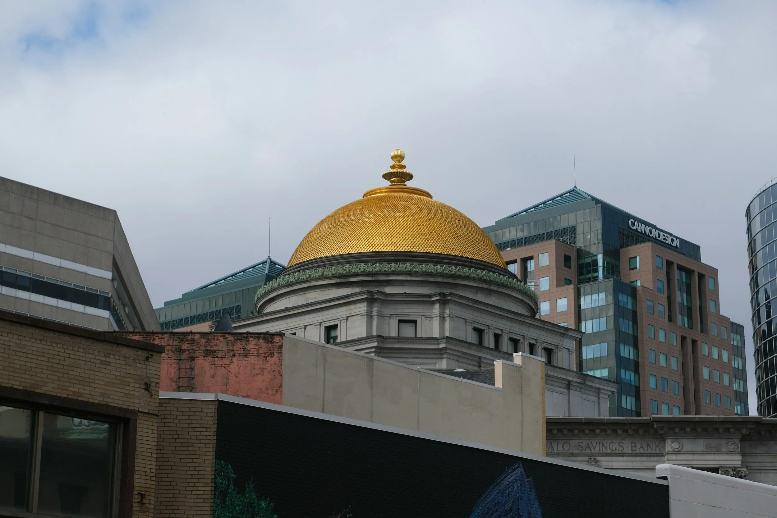 Cityscape featuring a building with a gold domed roof, surrounded by modern office buildings and older brick structures under cloudy sky.