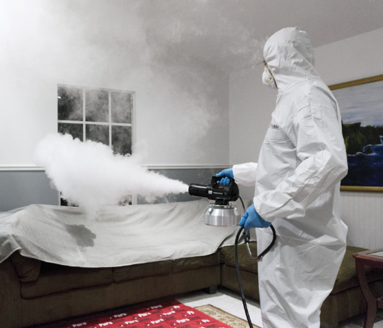 Person in protective suit using fogging machine to disinfect a living room.