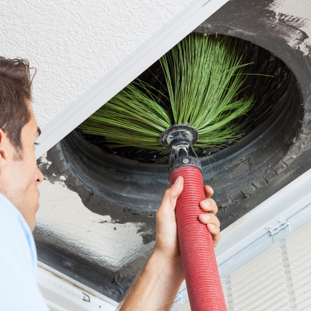 A person cleaning an air vent on the ceiling with a green brush attached to a red vacuum hose.