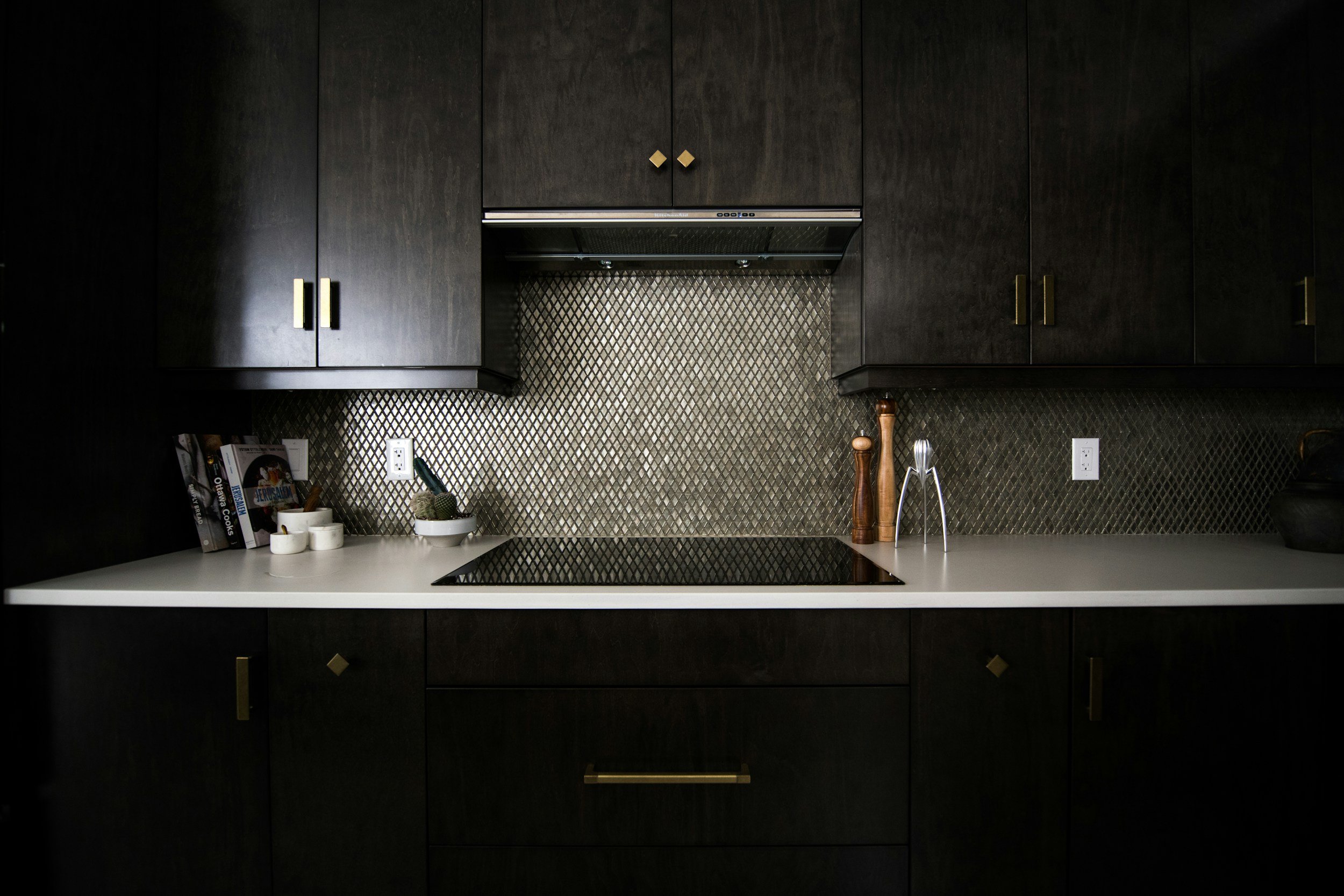 Dark kitchen cabinets with gold hardware, white countertop, black cooktop, and decorative items including books, small bowls, salt and pepper shakers, and a wall-mounted range hood on a textured gold backsplash.