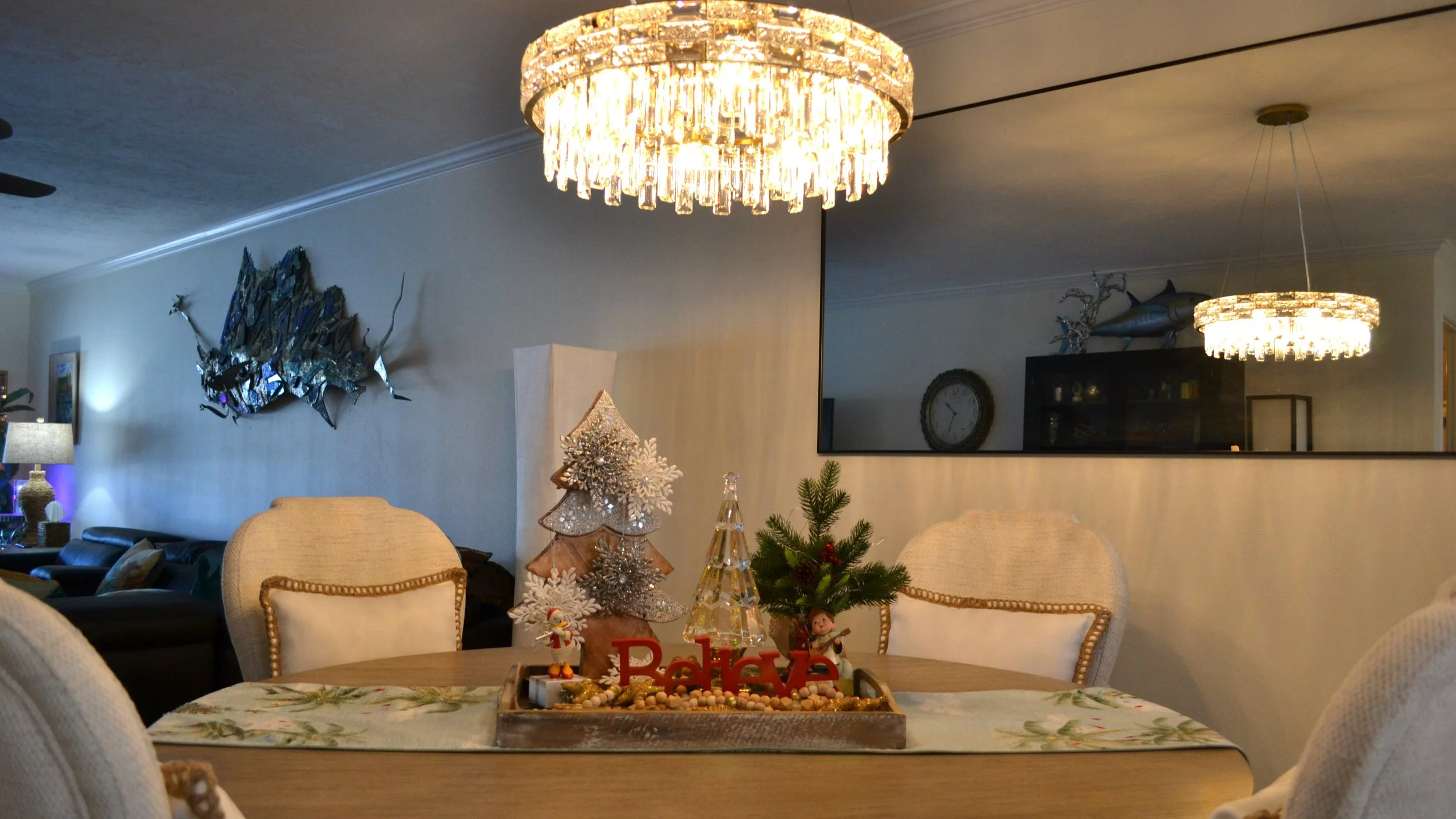 A dining room table with holiday decorations, including a small Christmas tree, a decorative wooden Christmas tree, and festive figurines. Two chandeliers hang from the ceiling, and the background features a wall with a mirror, a fish sculpture, and 