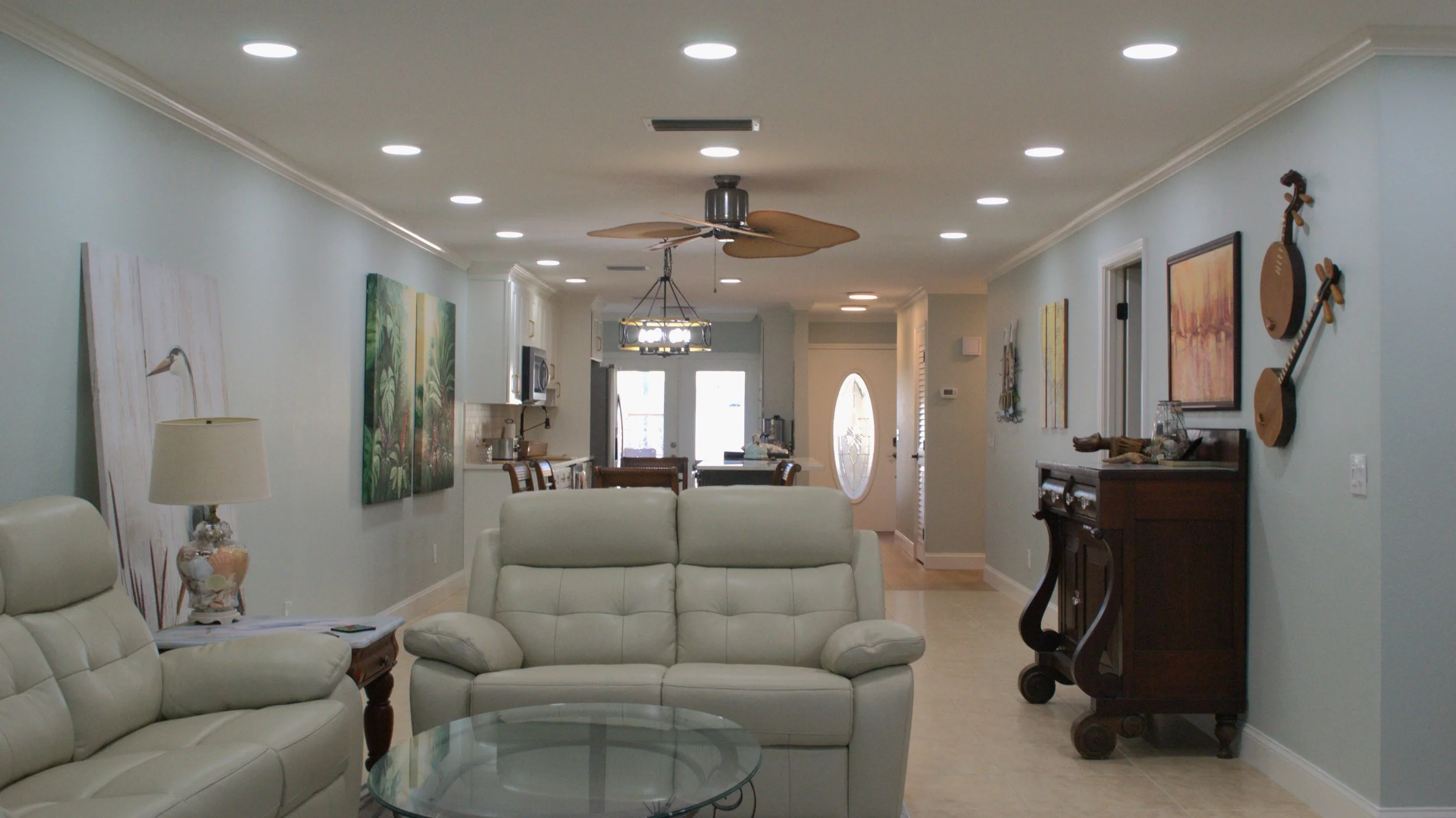 Living room with beige leather sofa and armchair, glass coffee table, side table with lamp, wall art, ceiling fan, and open view to dining and front entrance.