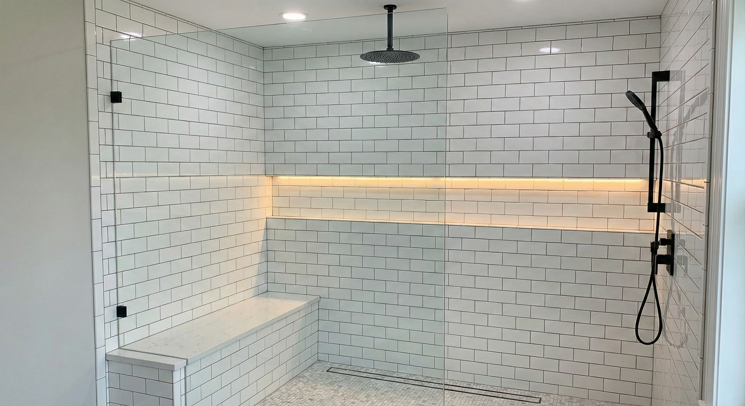 Modern walk-in shower with white subway tile walls, a built-in bench, a ceiling rain shower head, and a black handheld shower wand, with a glass shower enclosure and warm LED lighting.