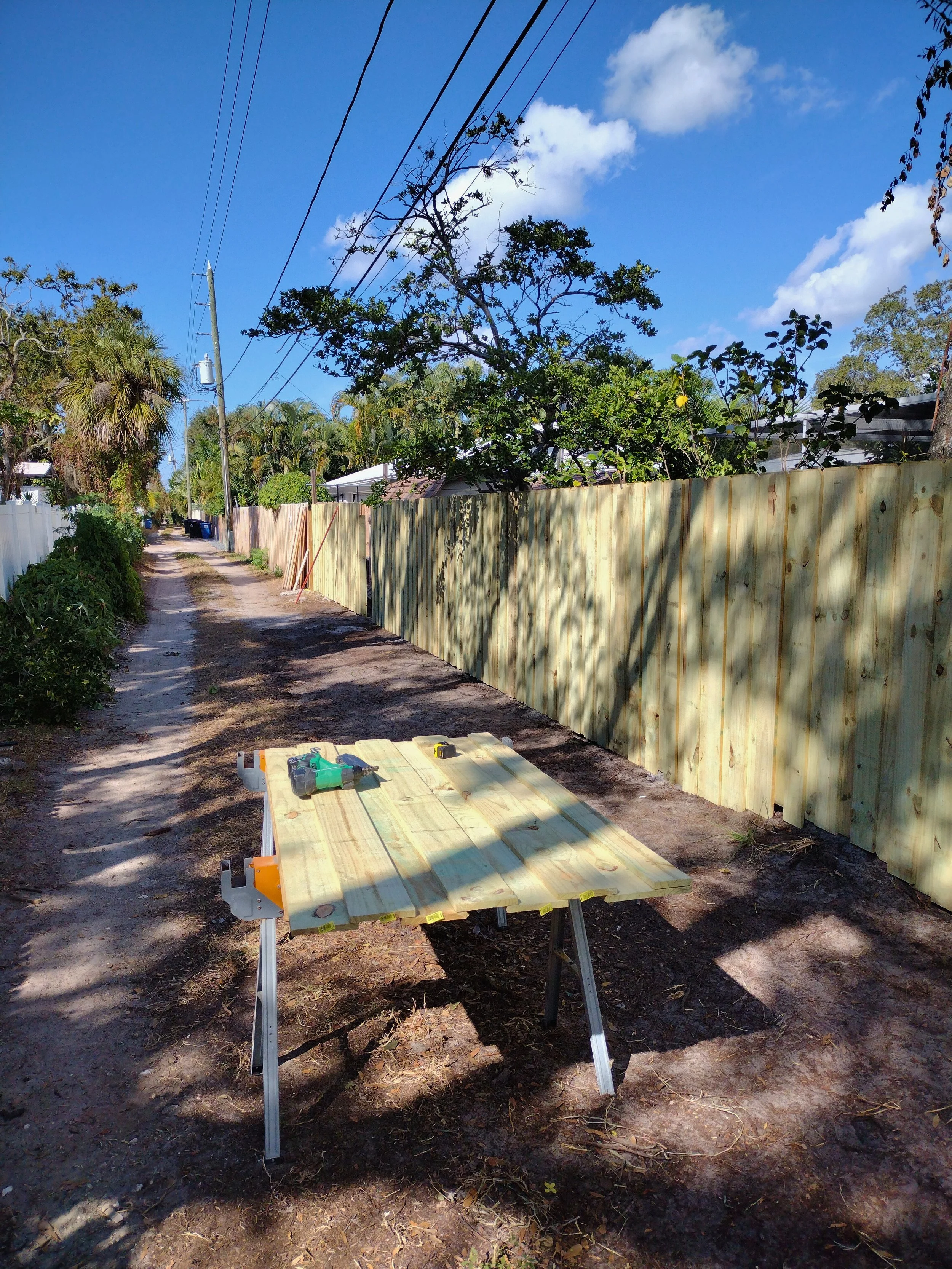 A construction site along a dirt road with a newly built wooden fence on the right and power lines overhead. A work table with tools is in the foreground, and trees and houses are visible in the background under a partly cloudy sky.