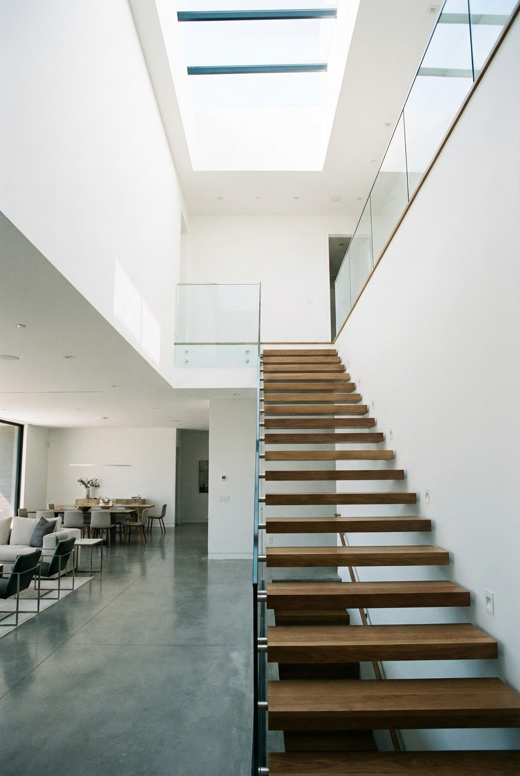 Modern interior with a staircase featuring wooden steps, a large skylight above, white walls, and a living area with chairs and dining table in the background.
