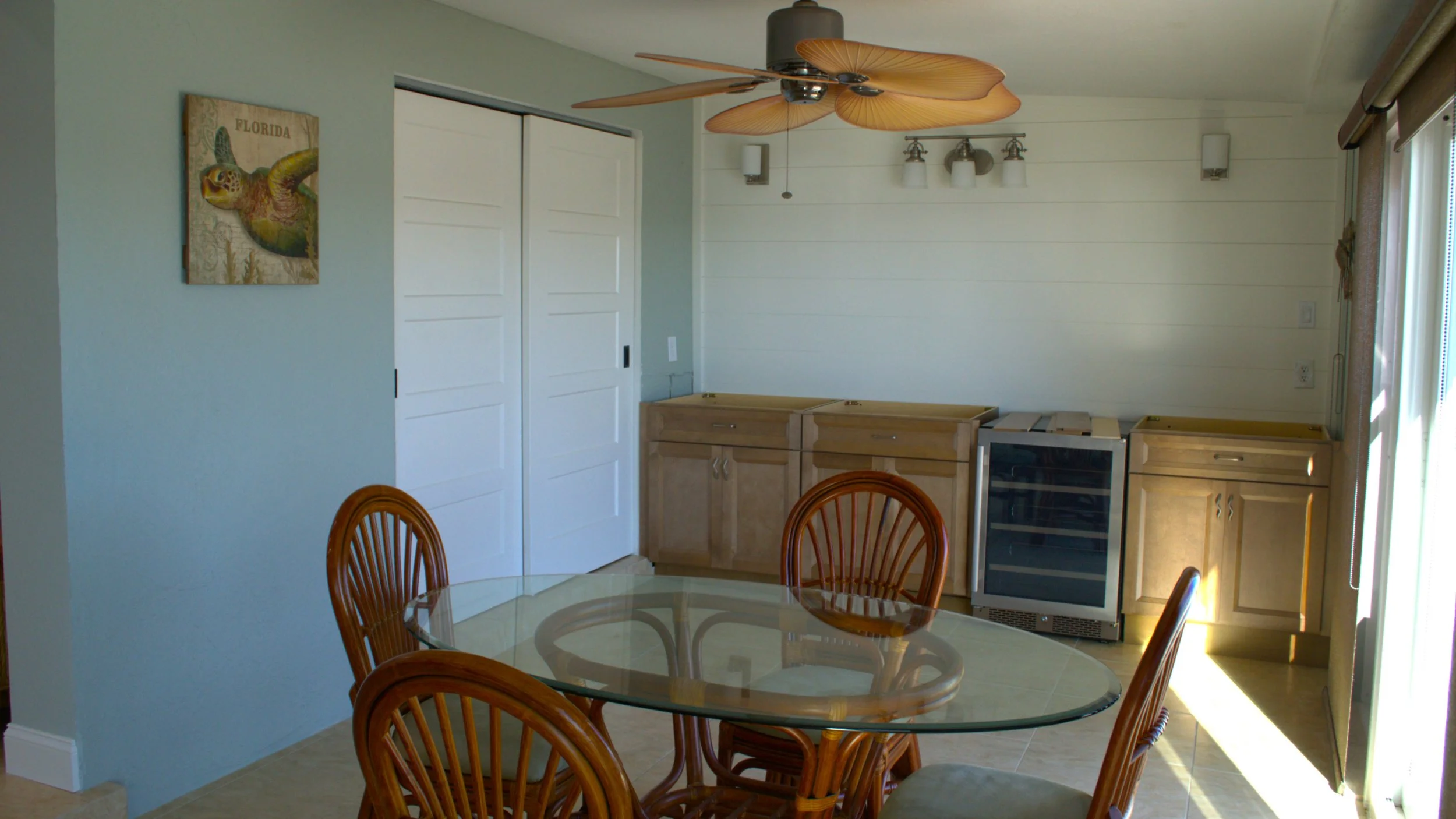 Empty kitchen area with a glass dining table, wooden chairs, light wood cabinets, a wine cooler, and a ceiling fan with leaf-shaped blades.