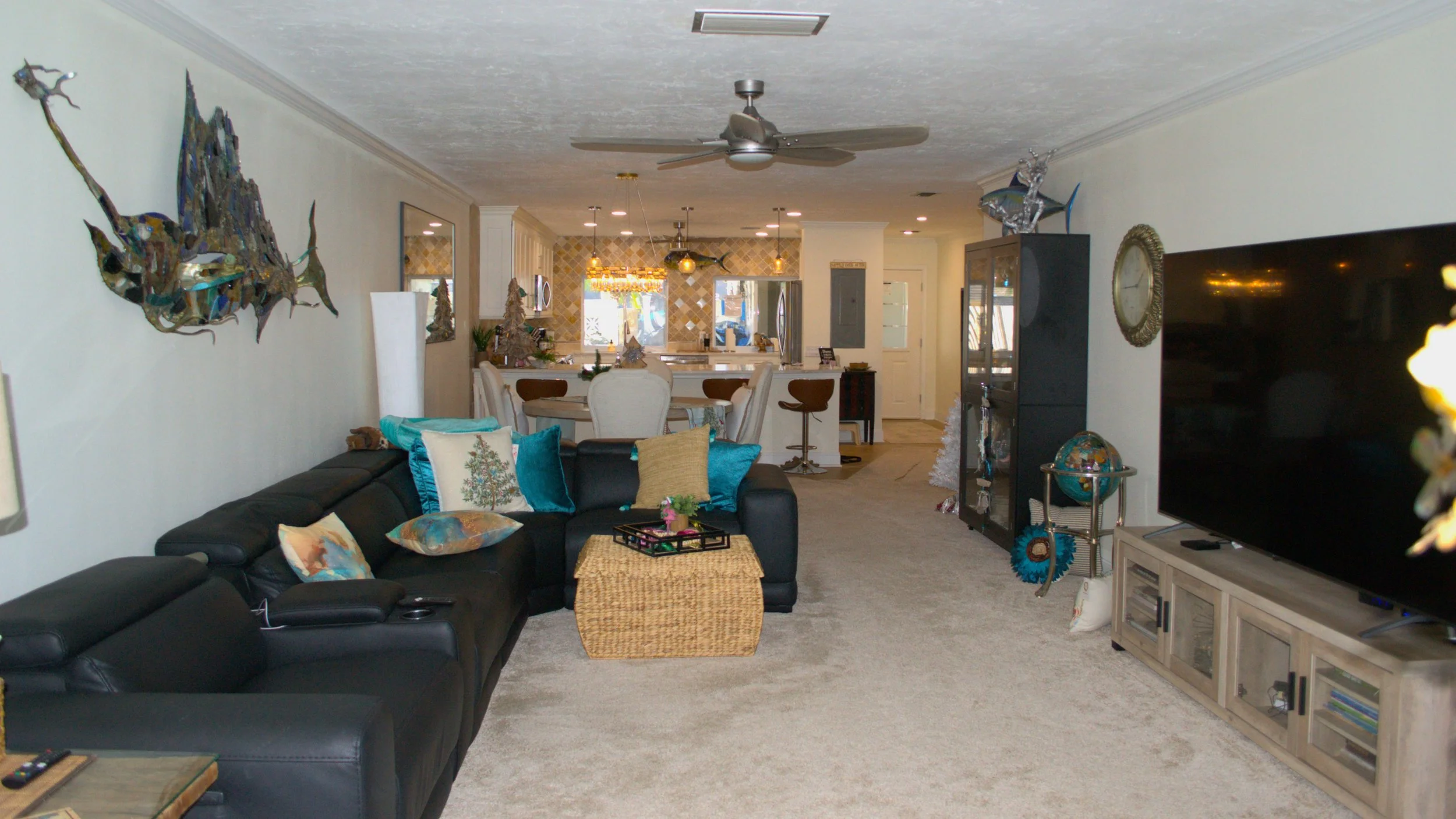 Living room with black leather sectional sofa, decorative pillows, and a wicker ottoman with tray and small plant. Wall art features a fish sculpture. TV on a light wood stand, globe, and shelving unit with decor. Open kitchen and dining area in back